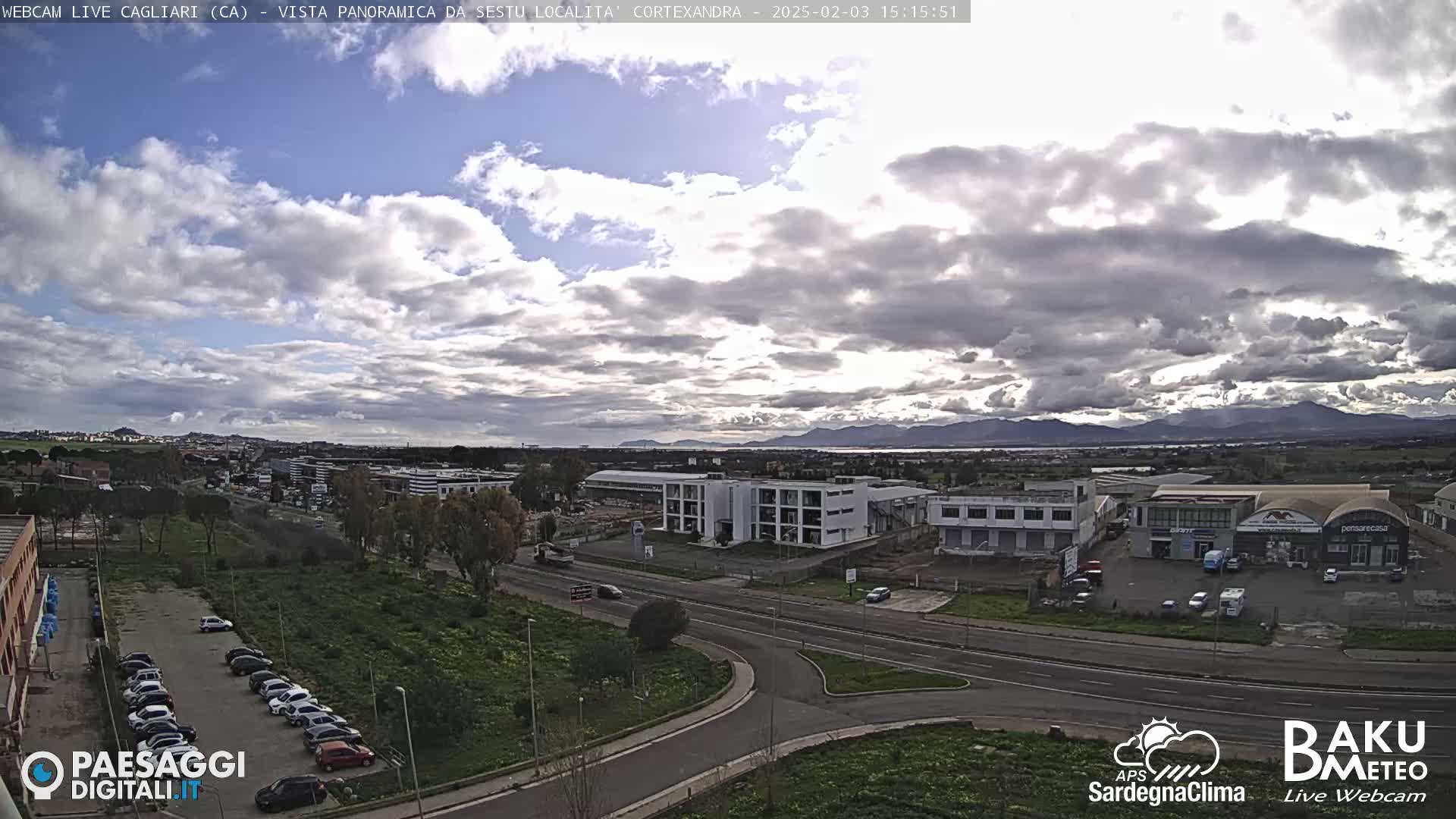 A partly cloudy day reveals a panoramic view of a city with industrial buildings and parking lots, a road system, and distant mountains under a mostly cloudy sky.