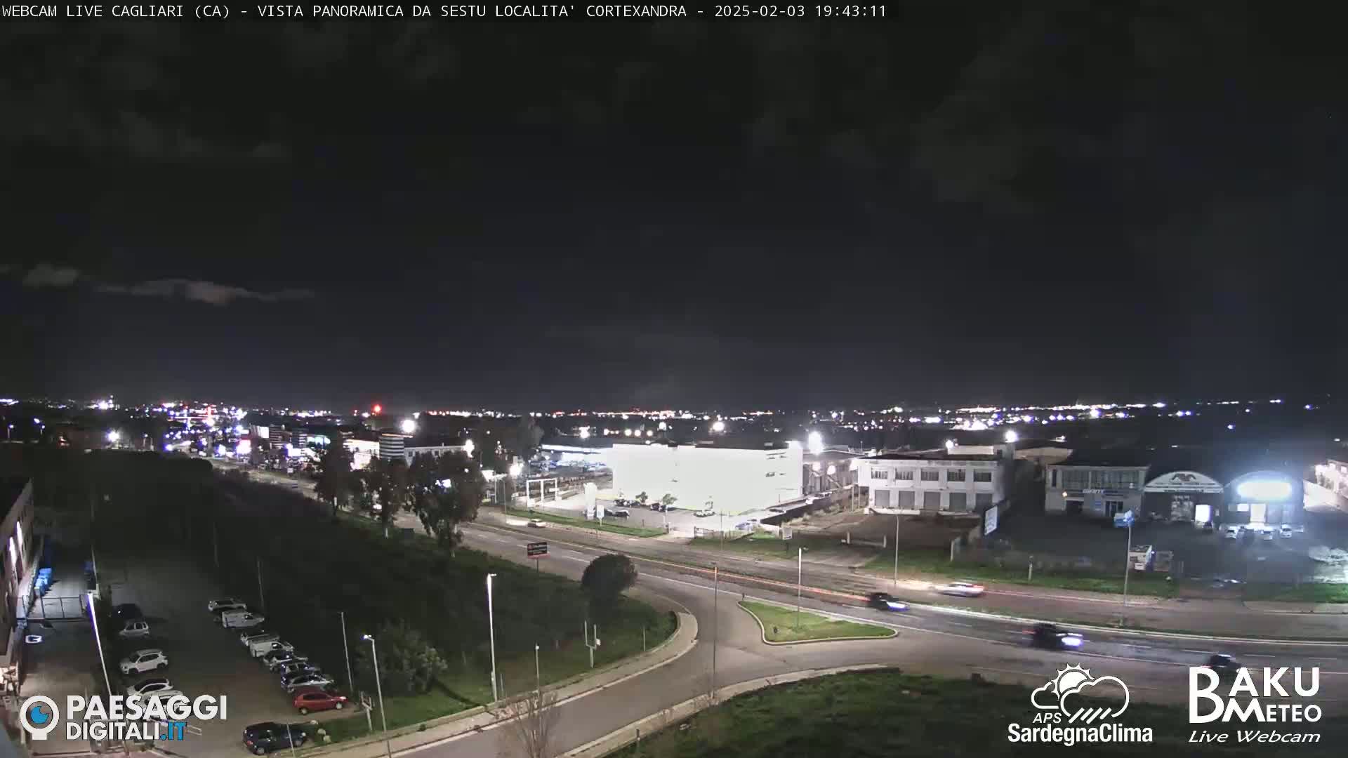 A nighttime, slightly cloudy view of a city's outskirts, showing roads, buildings, and parked cars.