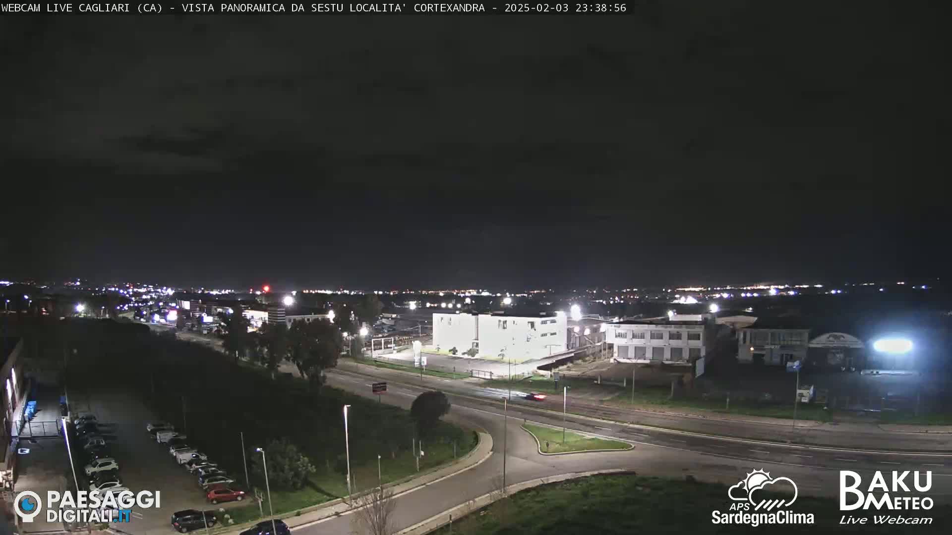 A nighttime, panoramic view of a city with roads, buildings, and parking lots under a dark, clear sky.