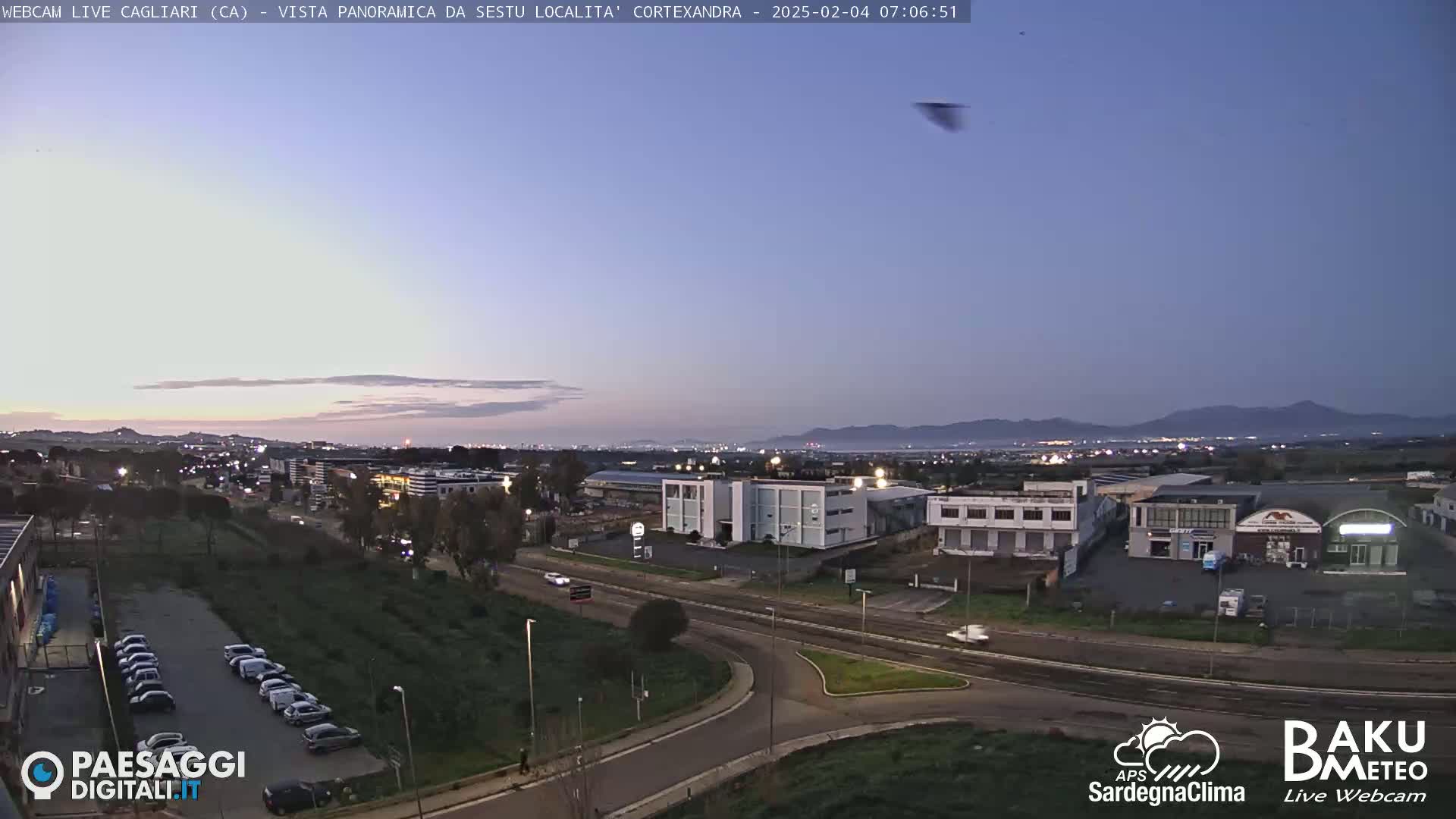 A twilight view of a road intersection near industrial buildings and a parking lot, with distant mountains visible under a clear, dusky sky.
