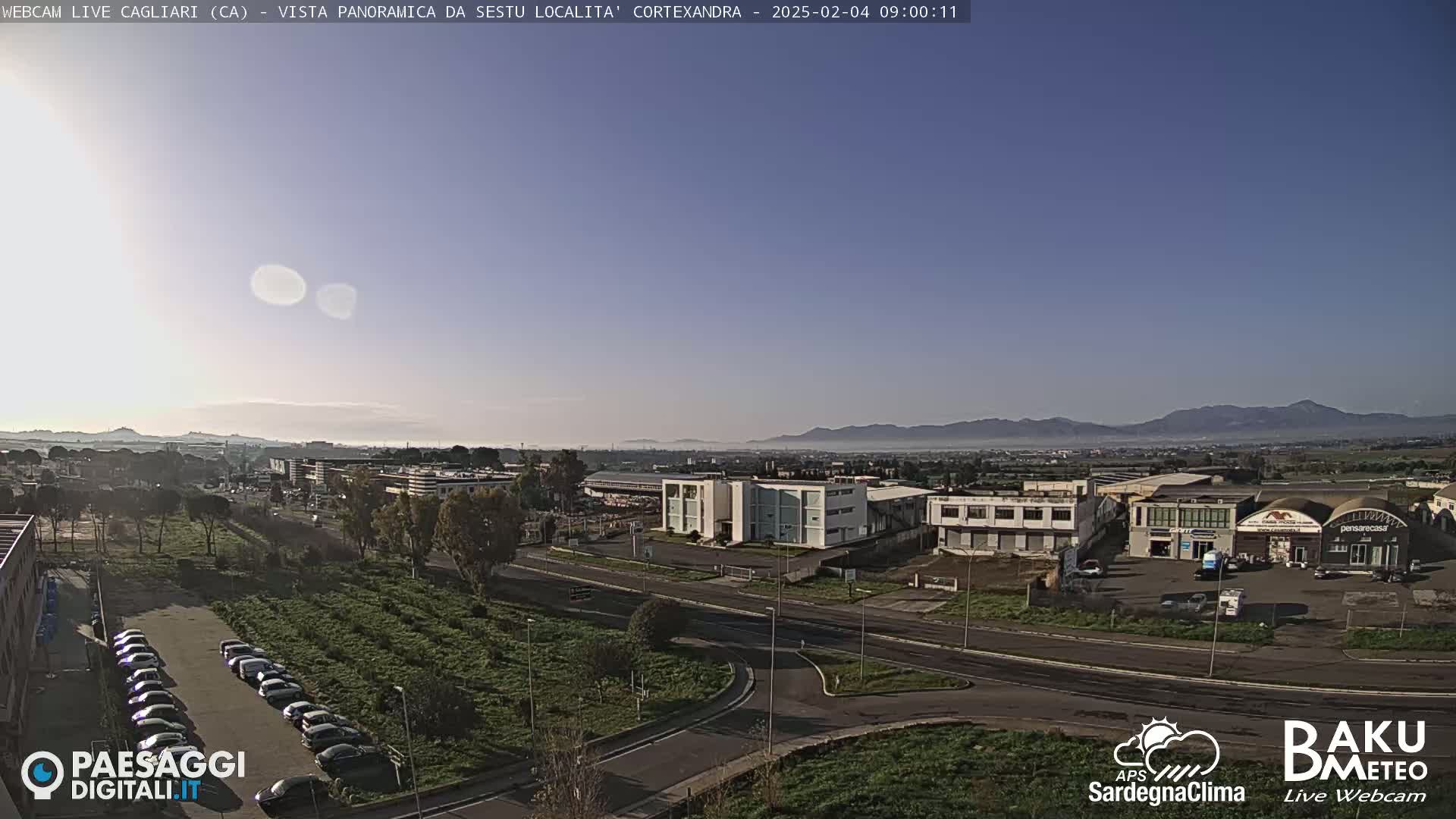 A clear, sunny day reveals a panoramic view of an industrial area and a parking lot, with distant mountains shrouded in a light haze in the background.