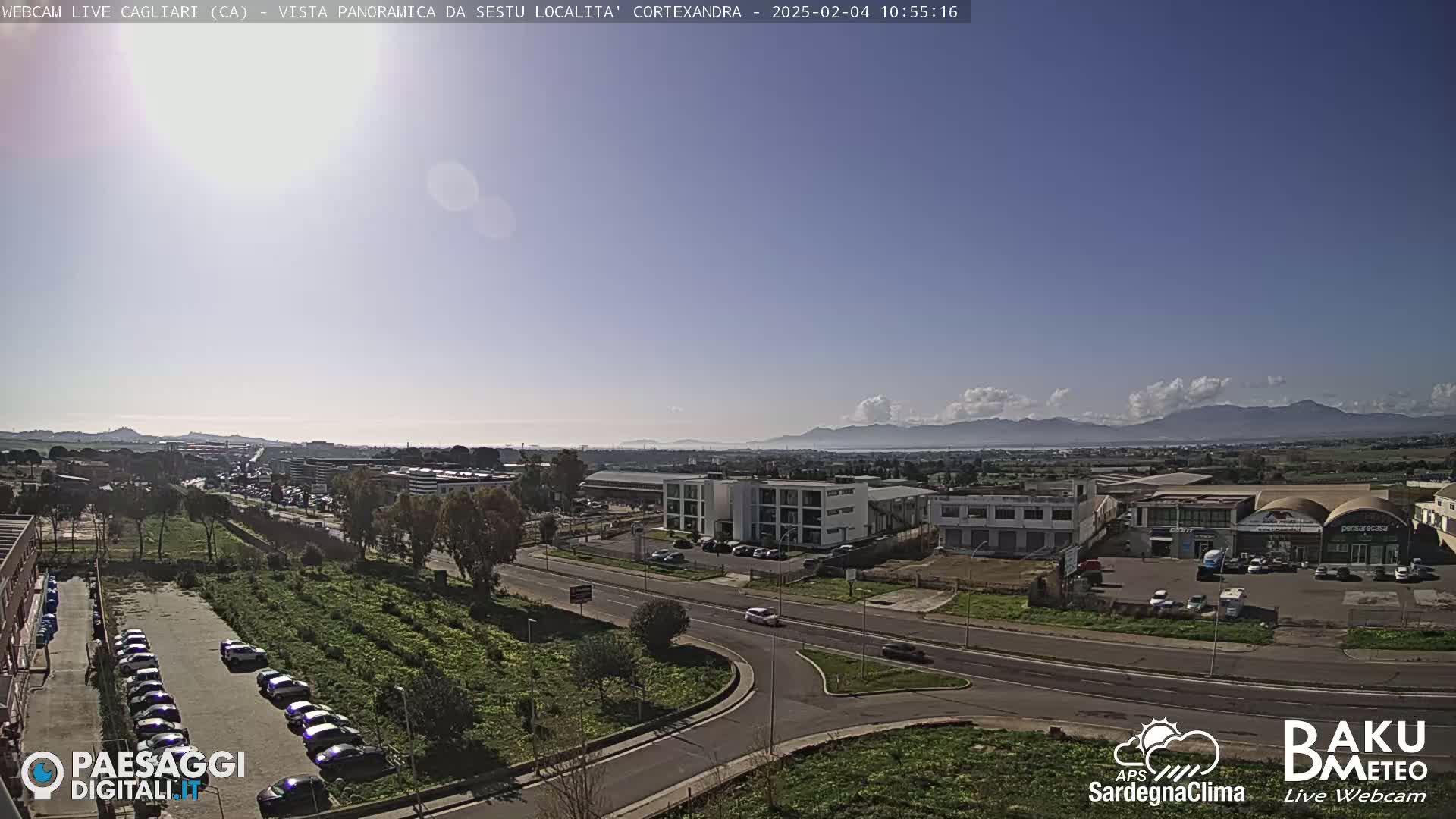 A sunny day reveals a panoramic view of a city's industrial area, roads, and parking lots, with distant mountains visible under a partly cloudy sky.