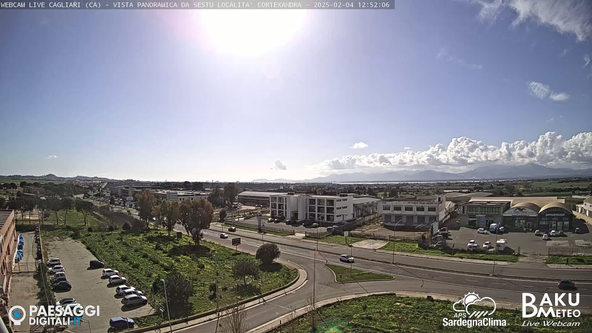 A sunny day with partly cloudy skies reveals a wide shot of a city landscape, featuring roads, buildings, and a parking lot, with distant mountains visible on the horizon.