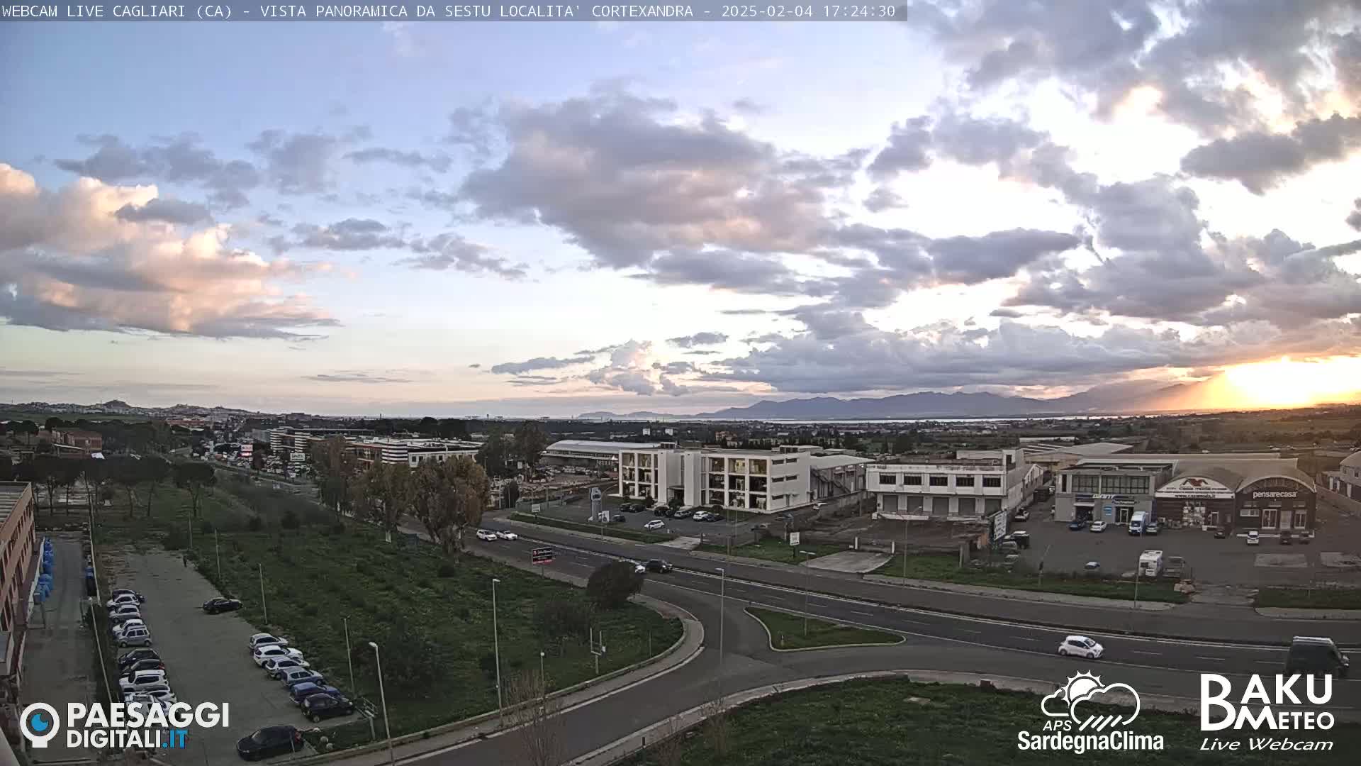 A partly cloudy sunset illuminates a cityscape with industrial buildings and roads, viewed from a high vantage point.