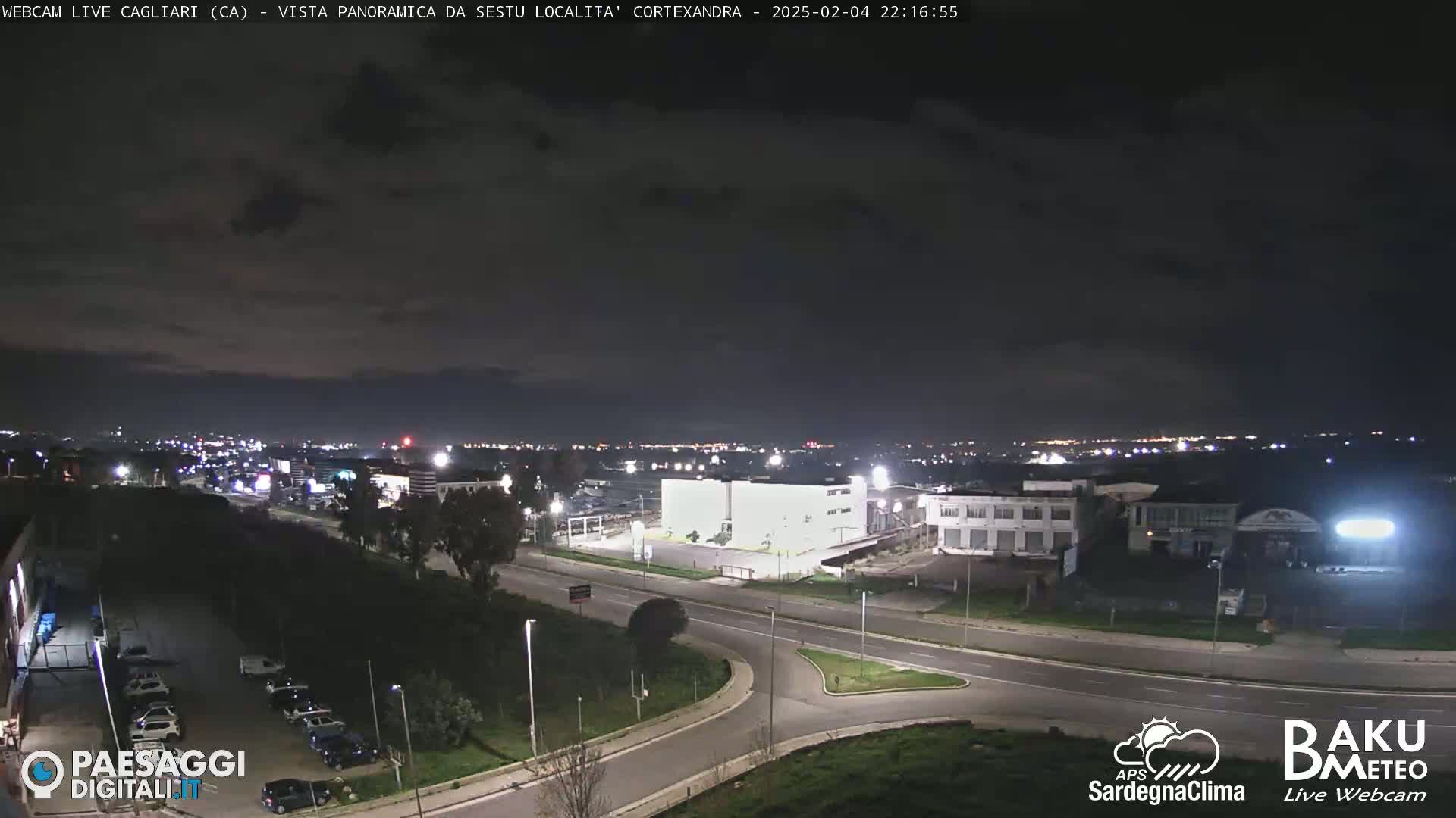 A nighttime, wide shot of a city road intersection and surrounding industrial area under a partly cloudy sky.