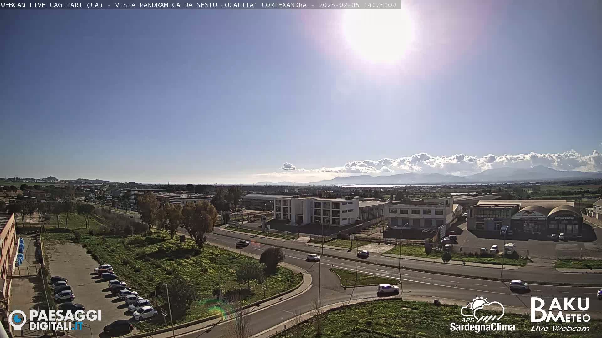 A sunny day reveals a panoramic view of a city with industrial areas, roads, and parked cars, backed by distant mountains with a layer of clouds along the horizon.