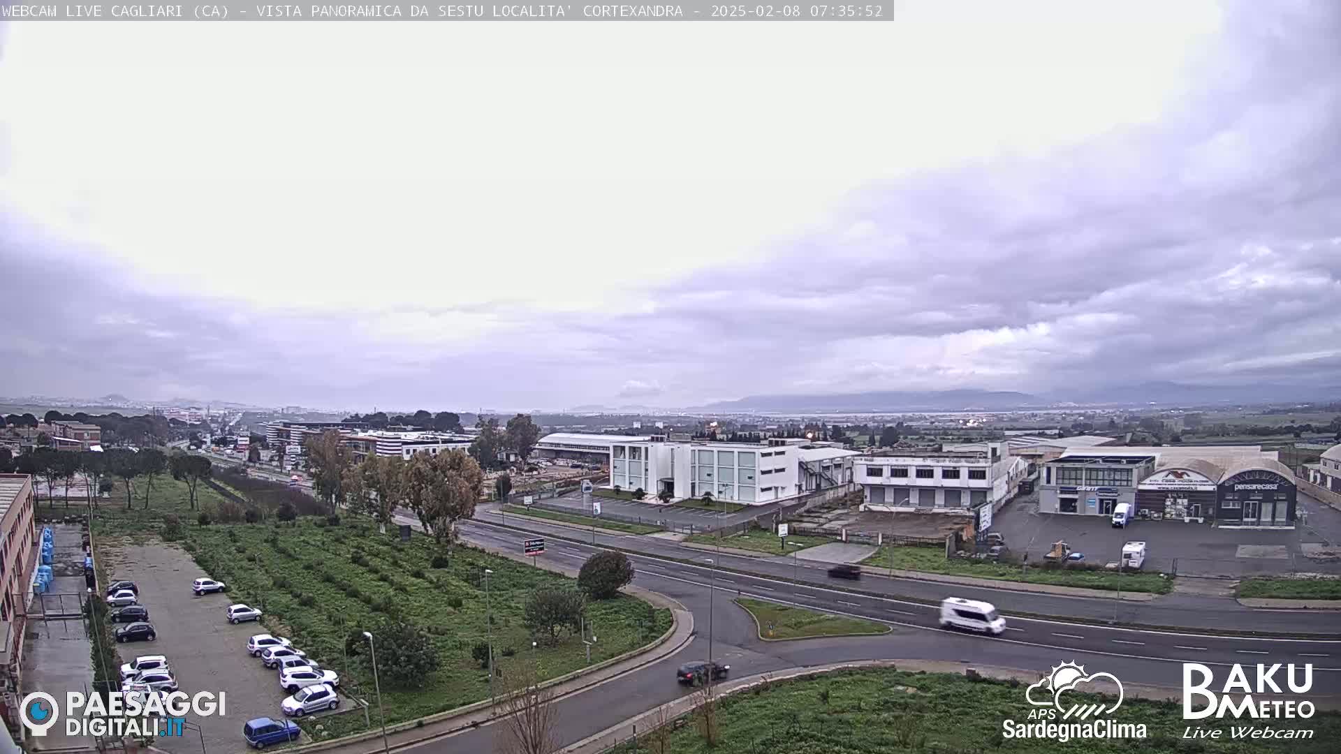 A wide, high-angle view of a road intersection and industrial area under an overcast sky, with cars parked in a lot and several vehicles traveling on the roads.