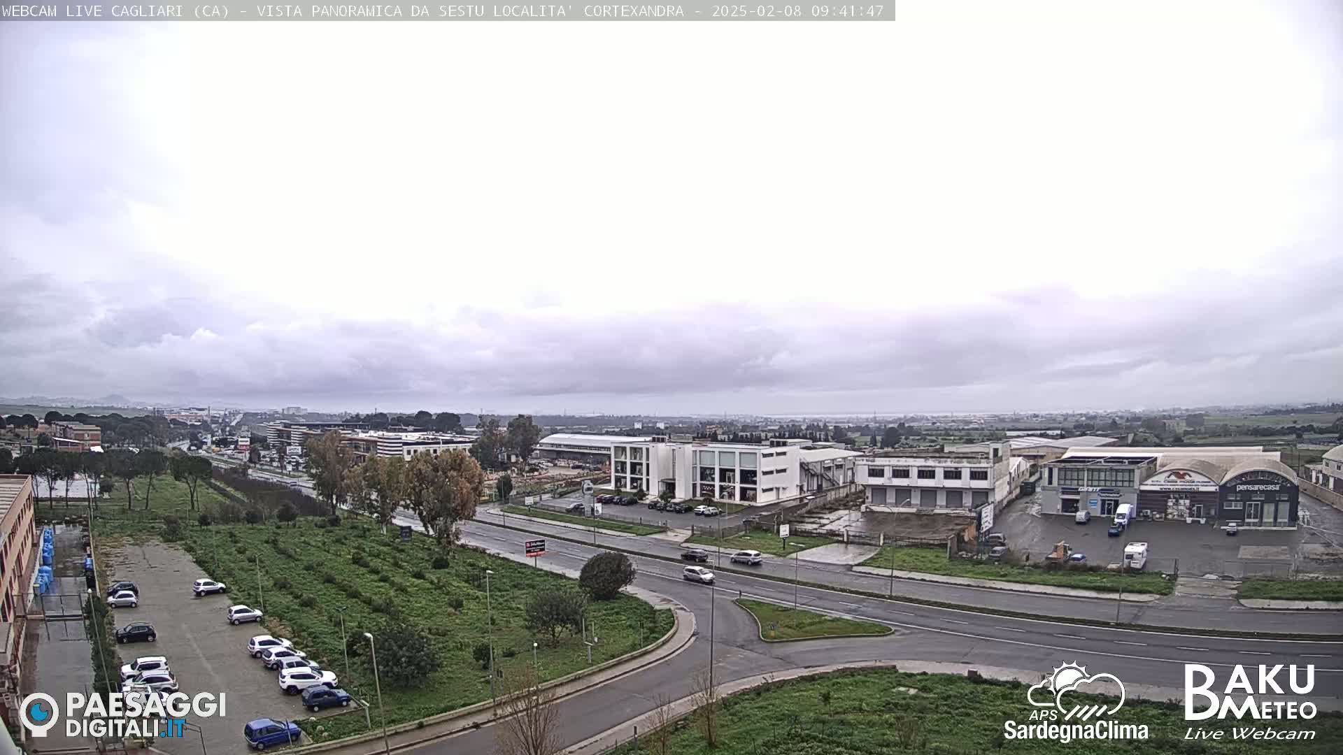 A wide, overcast view of a city's industrial and residential areas, with a road and parking lots visible, and a few cars driving by.