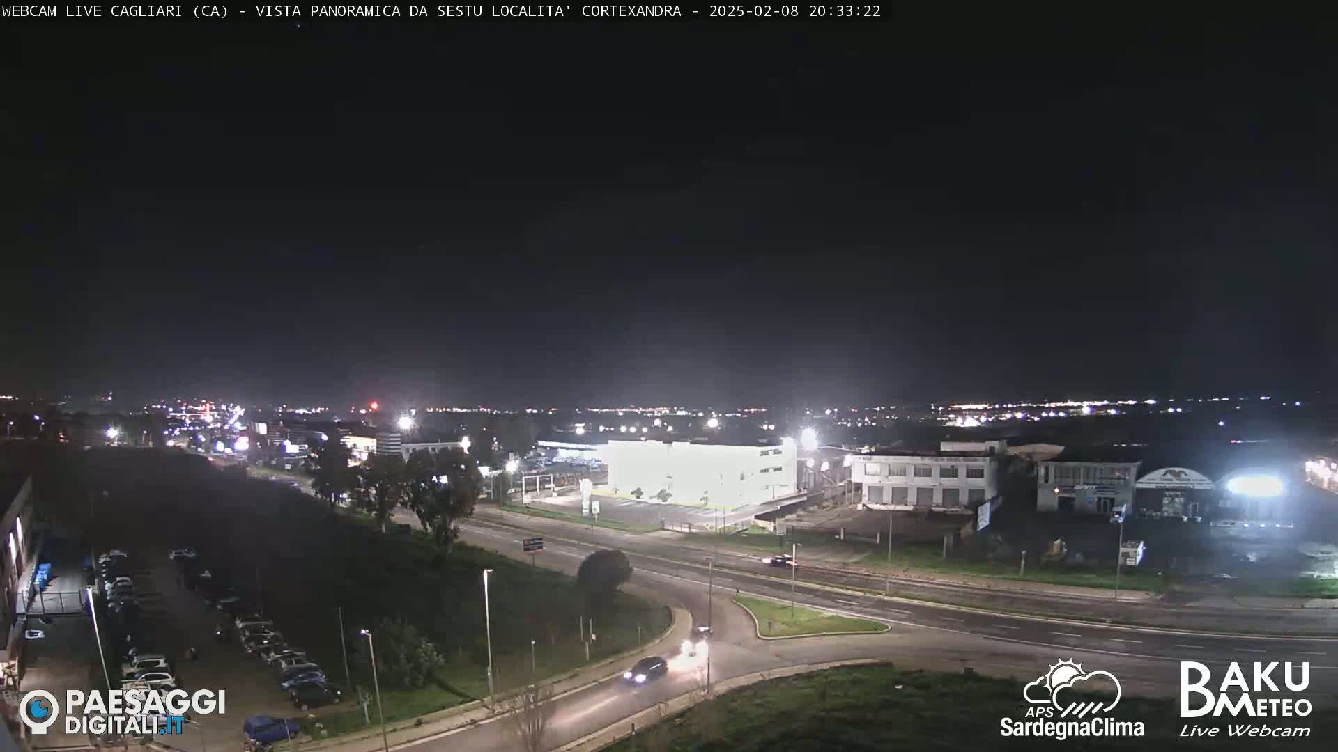 A nighttime, panoramic view of a city's outskirts, showing roads, buildings, and parking lots under clear skies.