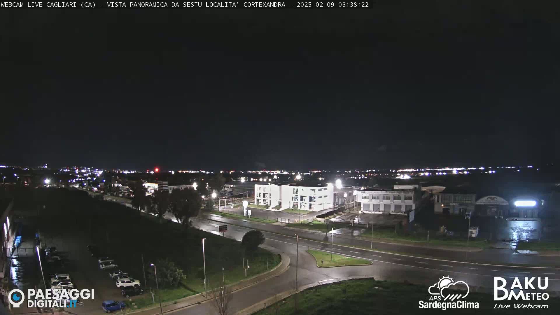 A nighttime, wide shot of a city's outskirts, featuring roadways, buildings, and some parked cars under a dark sky.