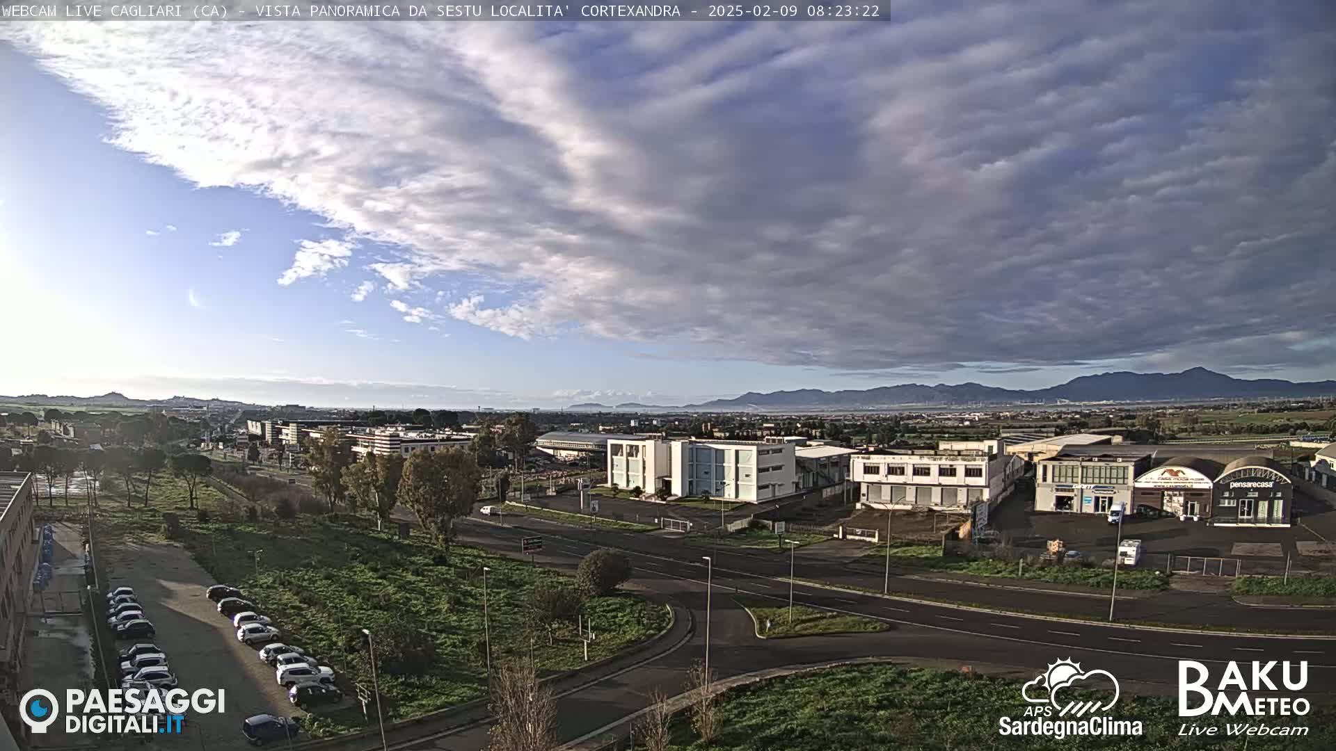 A mostly cloudy sky hangs over a flat landscape of buildings and roads, with distant mountains visible on the horizon.