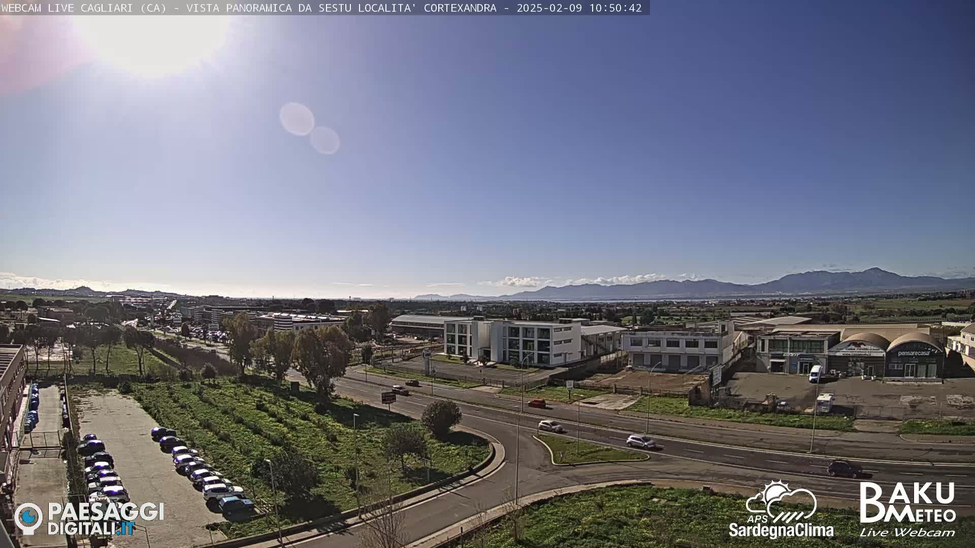 A clear, sunny day reveals a panoramic view of a cityscape with industrial buildings, roads, and a parking lot, bordered by green spaces and mountains in the distance.