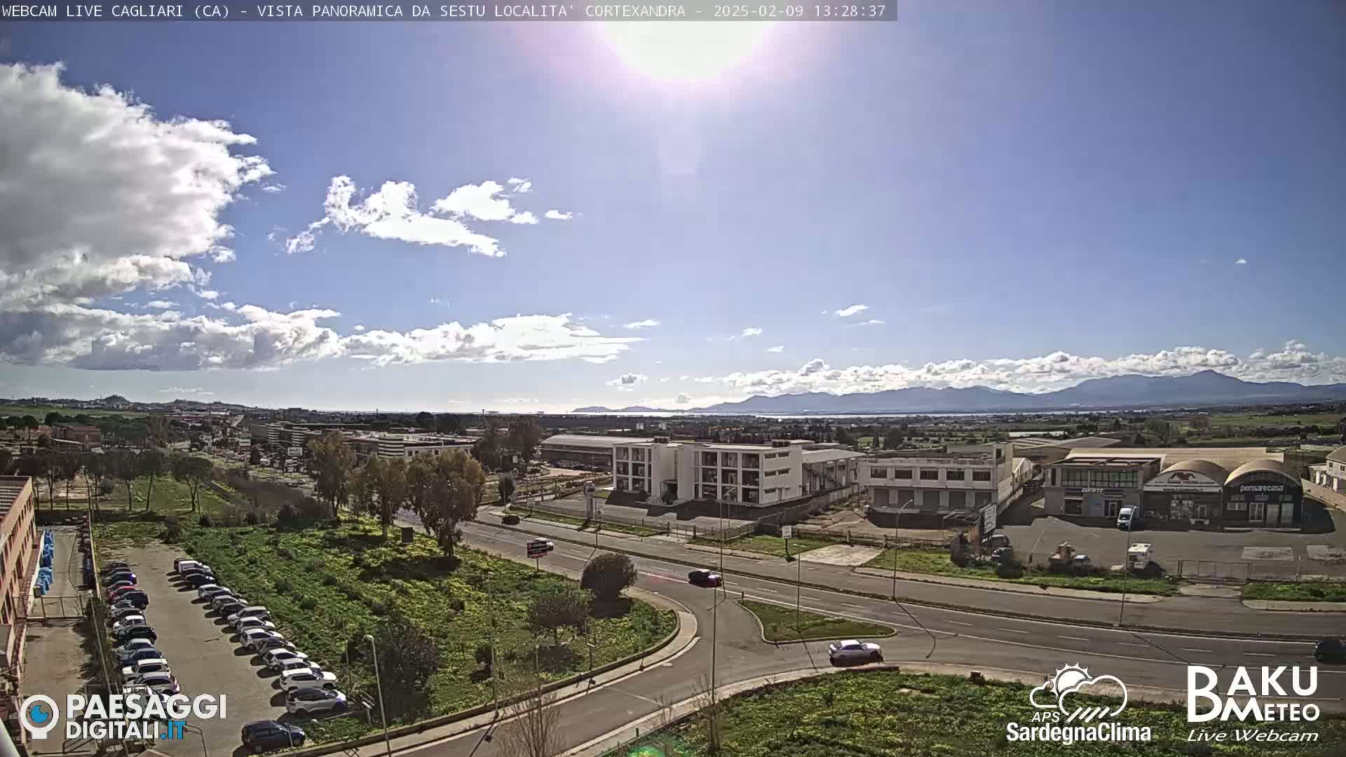 A partly cloudy day reveals a panoramic view of a city with industrial buildings, roads, and parked cars, backed by distant mountains and a body of water.