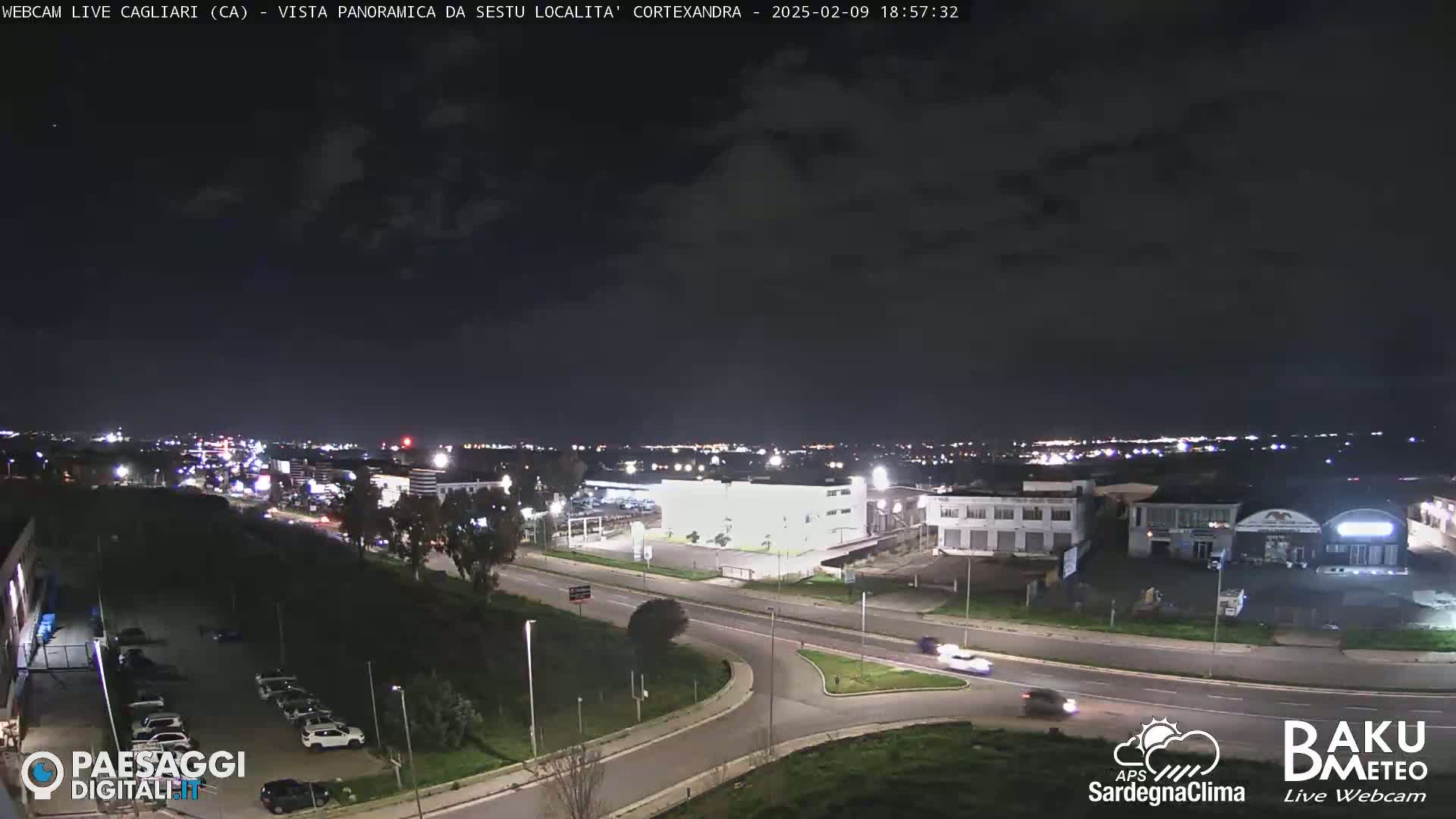 A nighttime, mostly cloudy view of a city with roads, buildings, and cars, illuminated by streetlights.