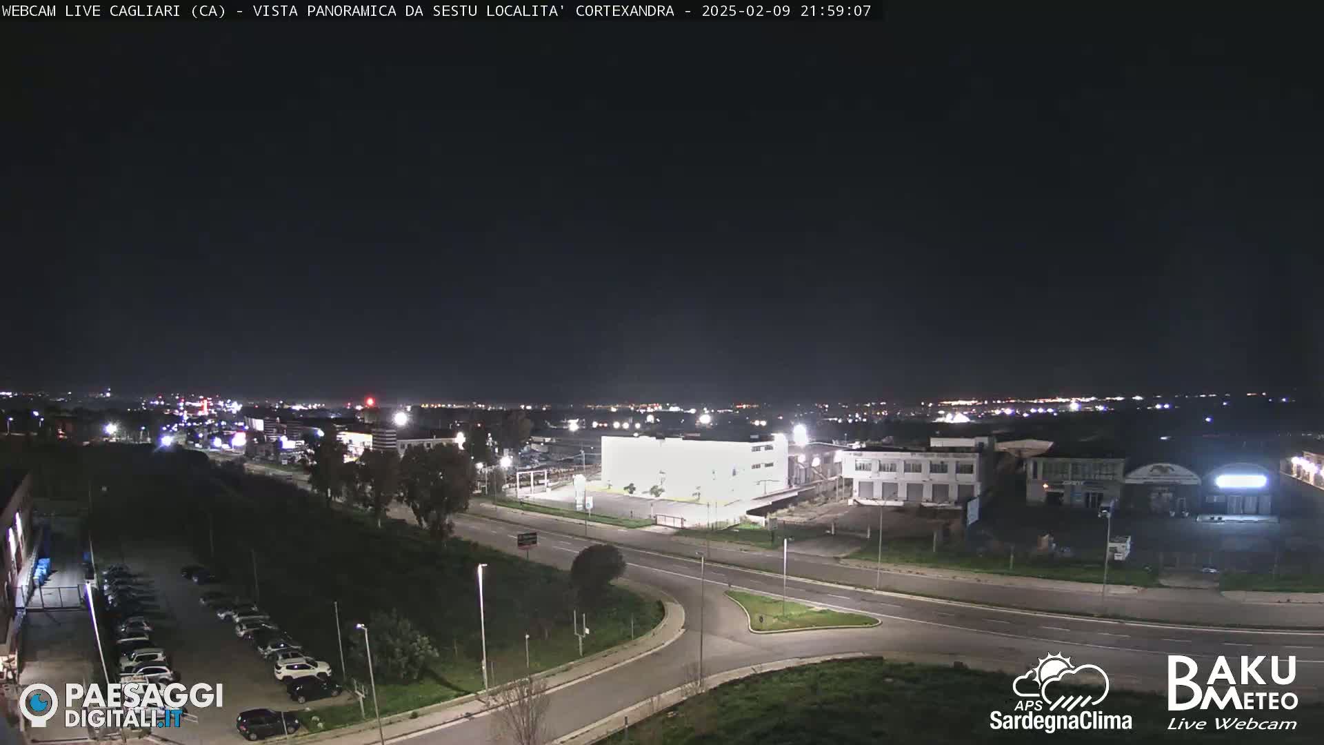 A nighttime, panoramic view of a city featuring roads, buildings, and parking lots under a clear, dark sky.
