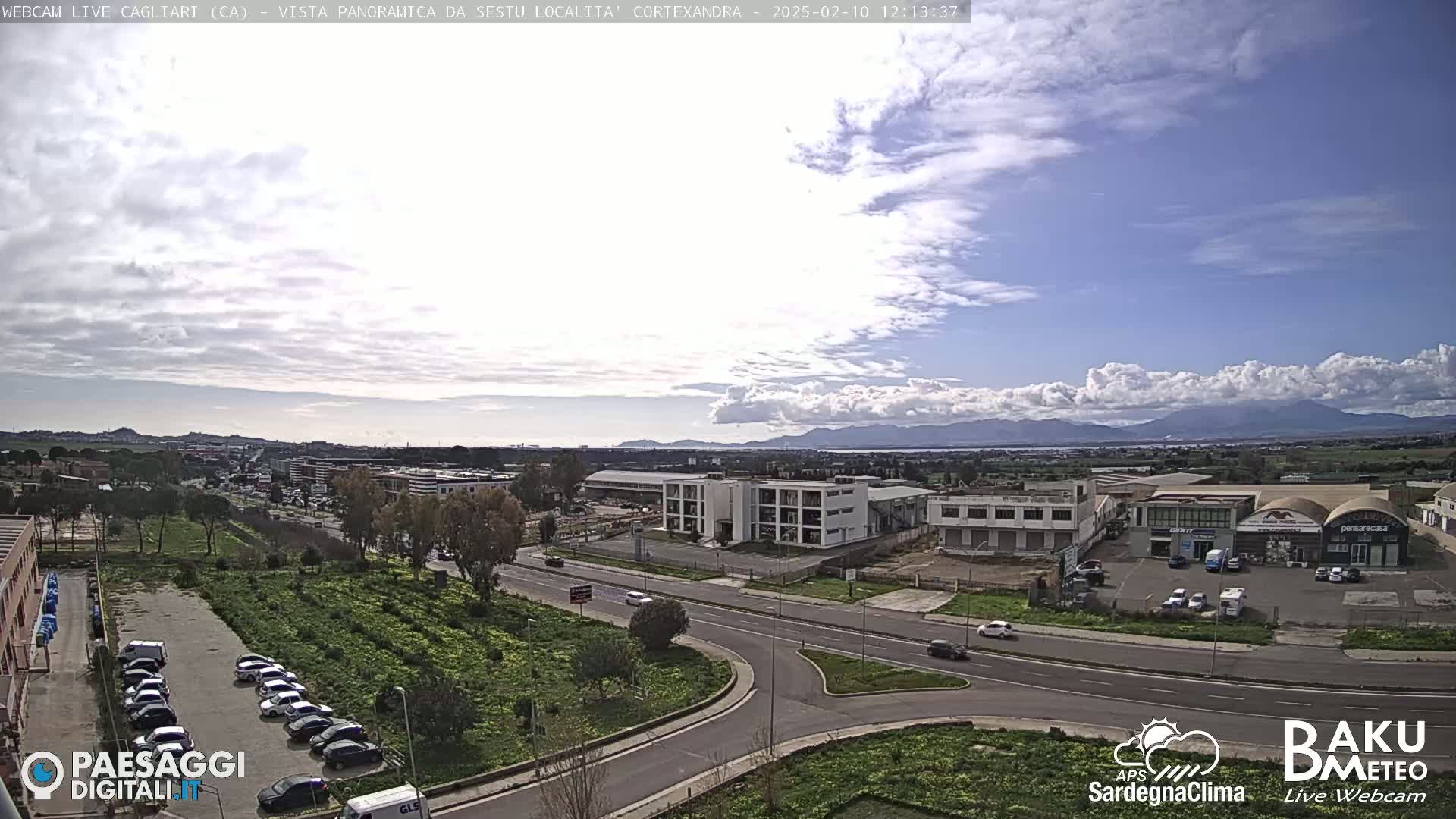 A partly cloudy day reveals a panoramic view of a city with industrial buildings, roads, and parked cars, mountains in the distance.