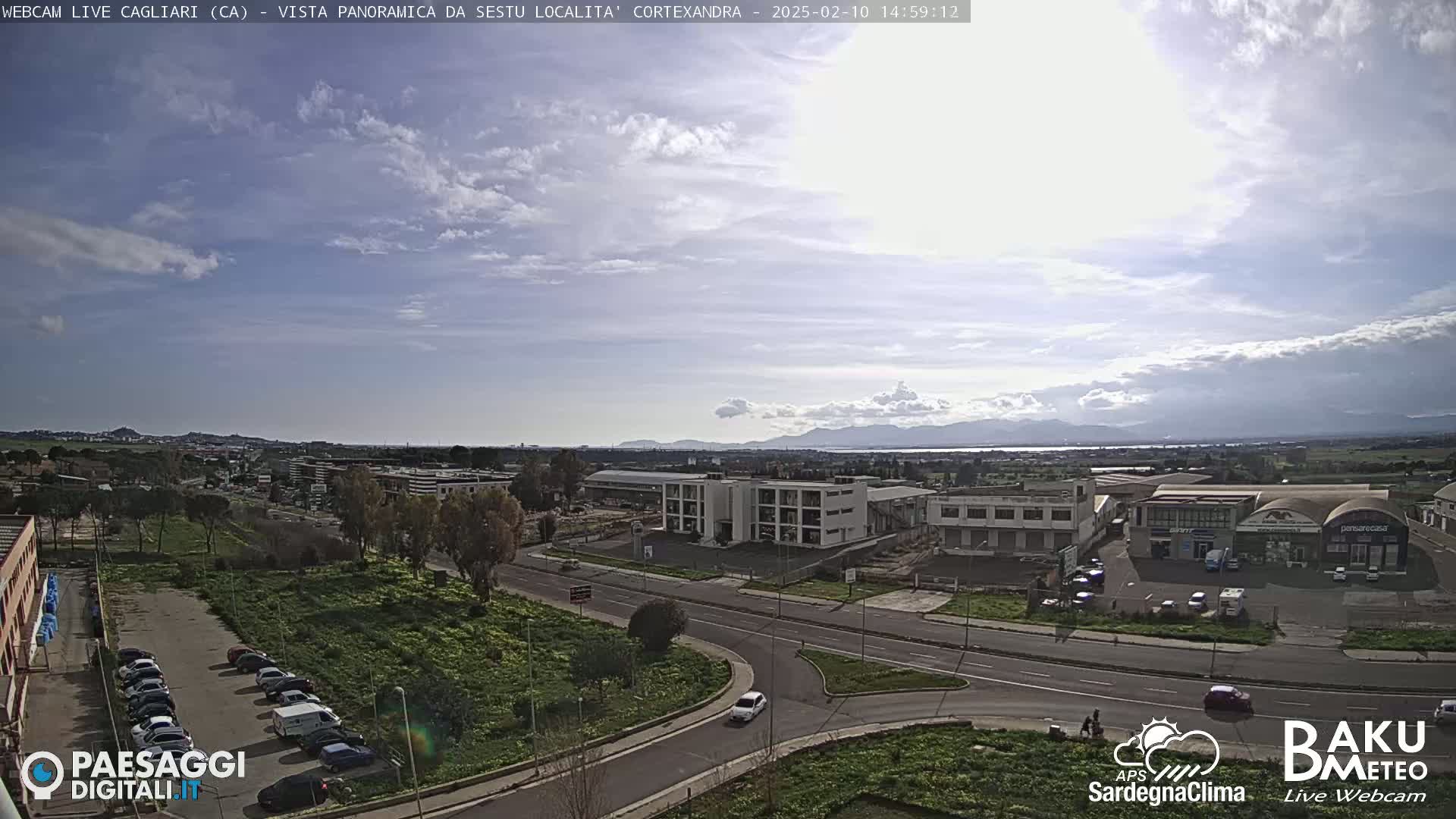 A mostly sunny day reveals a panoramic view of a city with industrial buildings and roadways, mountains in the distance, and a parking lot with several cars.