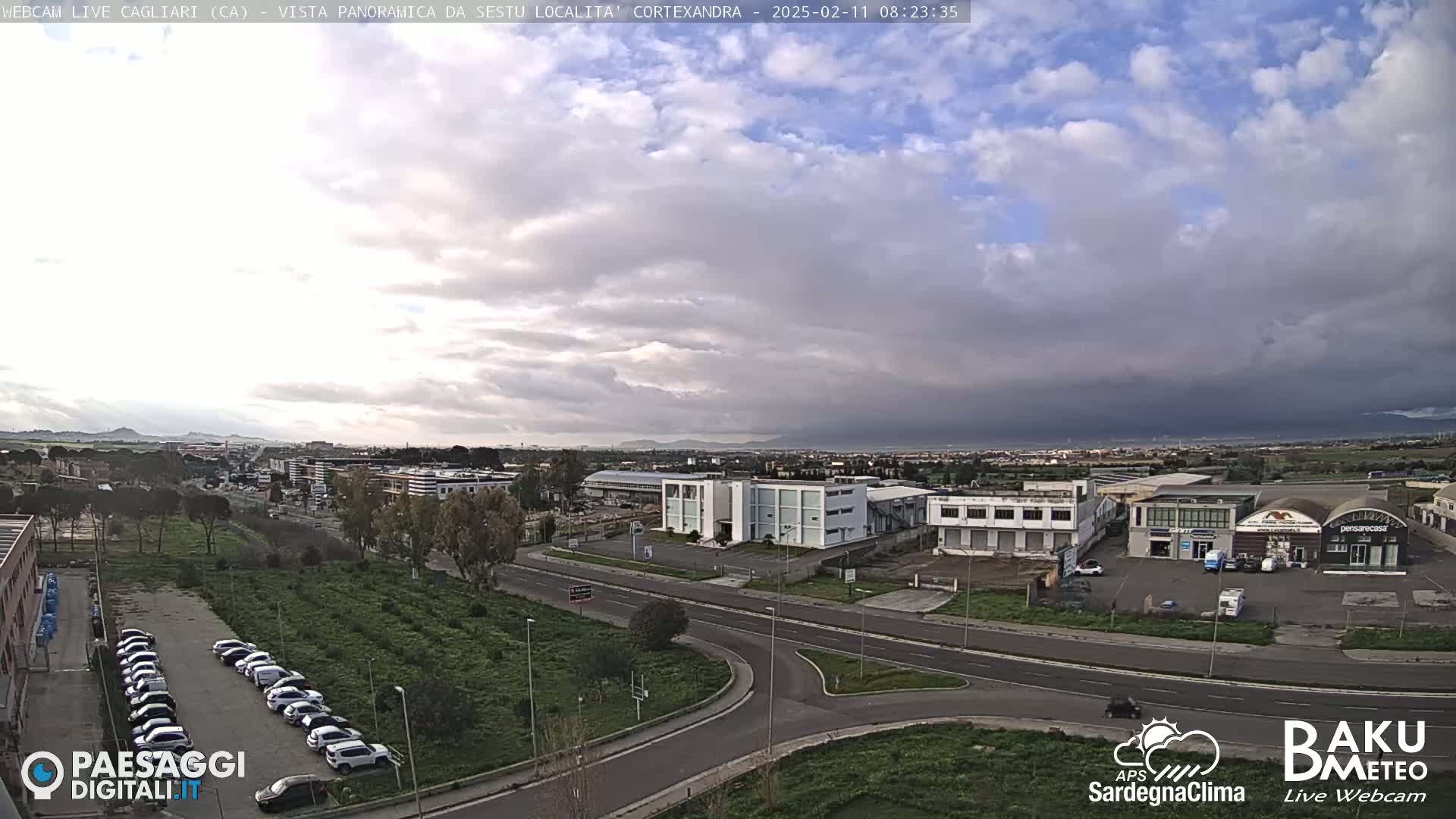 A mostly cloudy, overcast day reveals a wide shot of an industrial area with roads, buildings, and parked cars, extending to a distant mountainous horizon.