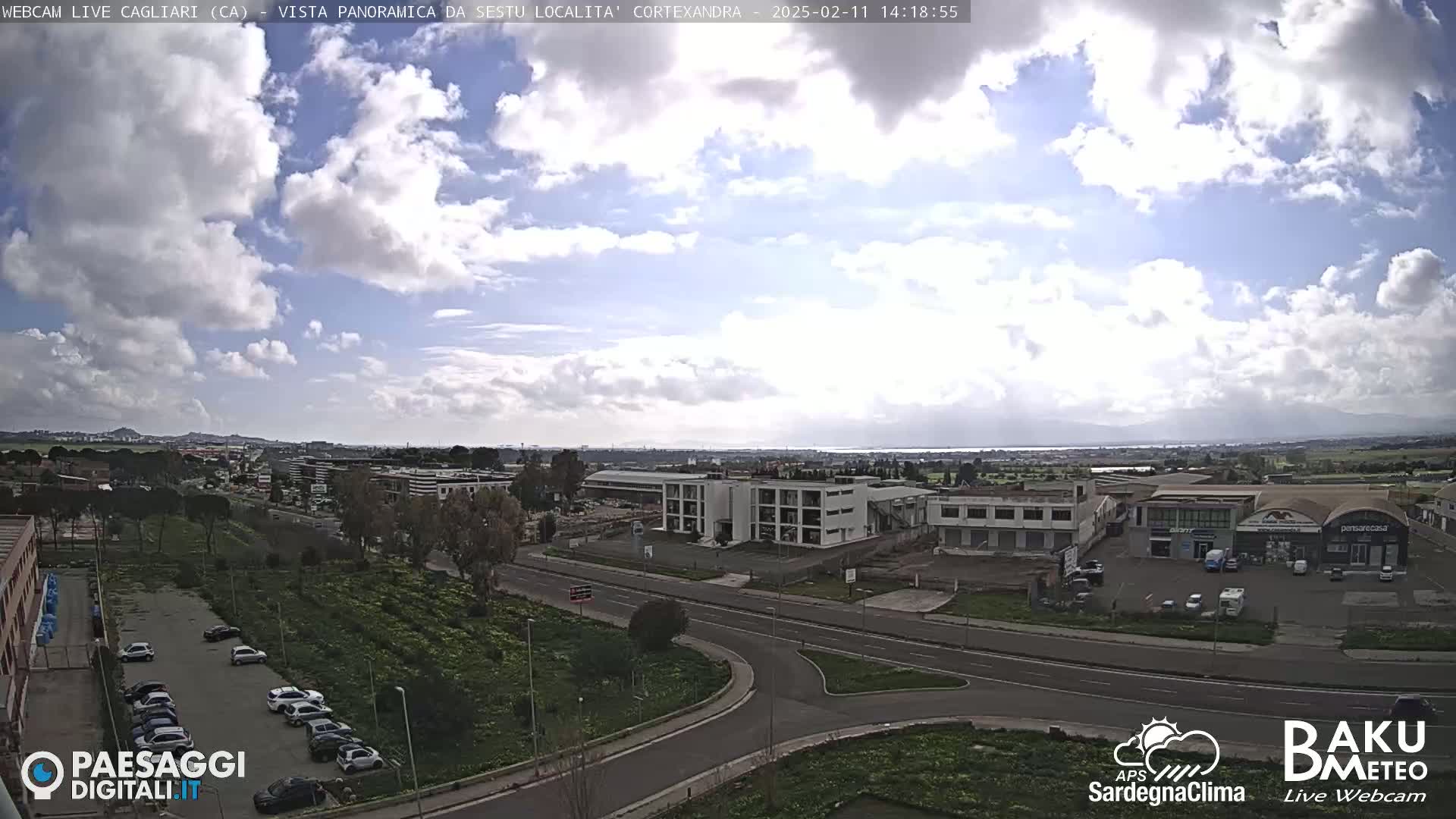 A partly cloudy day reveals a panoramic view of a city with roads, buildings, and a parking lot, extending to a body of water in the distance.