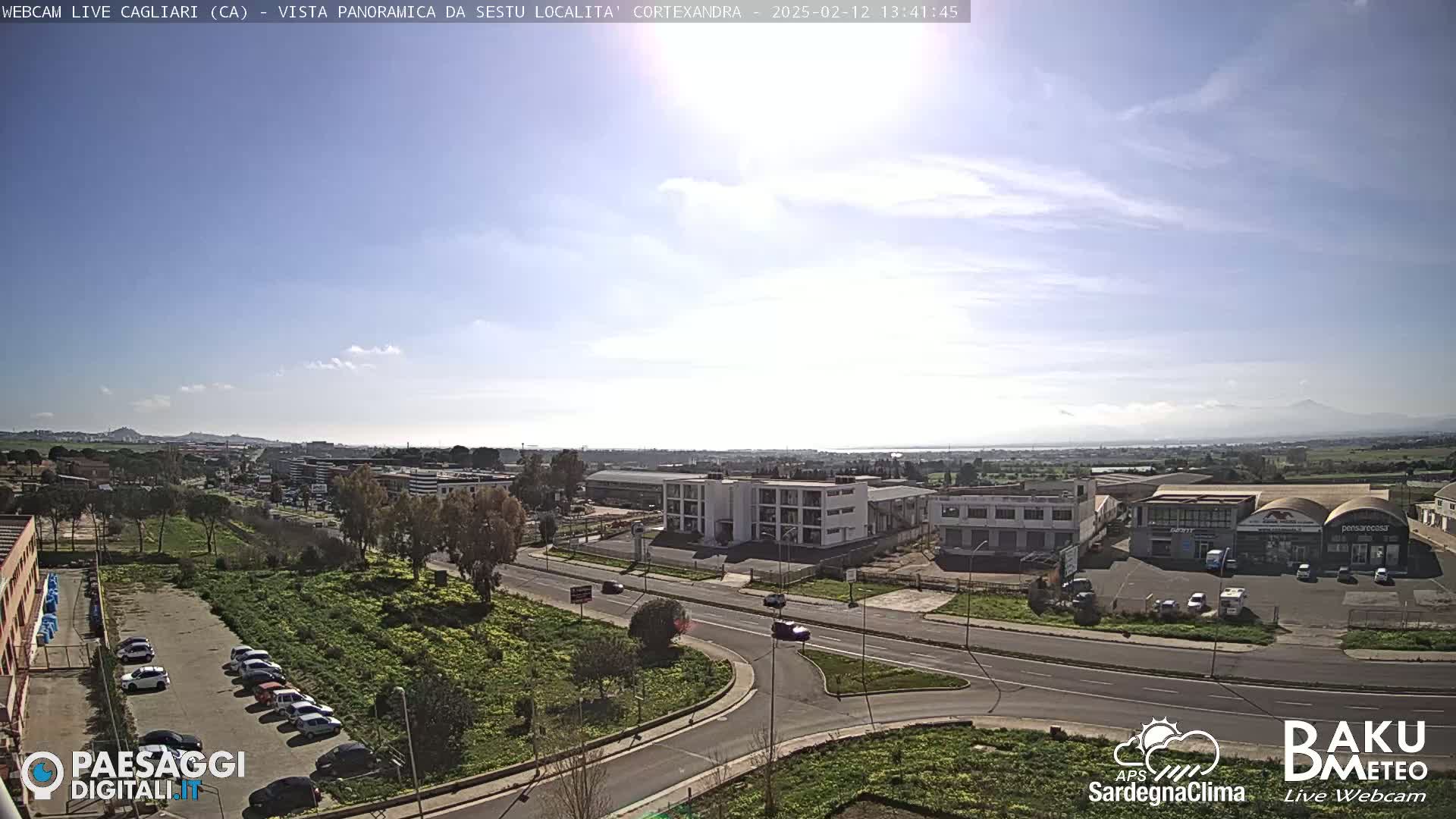 A sunny day reveals a wide shot of a city's outskirts, featuring industrial buildings, roads, and parked cars, with mountains visible in the distance.