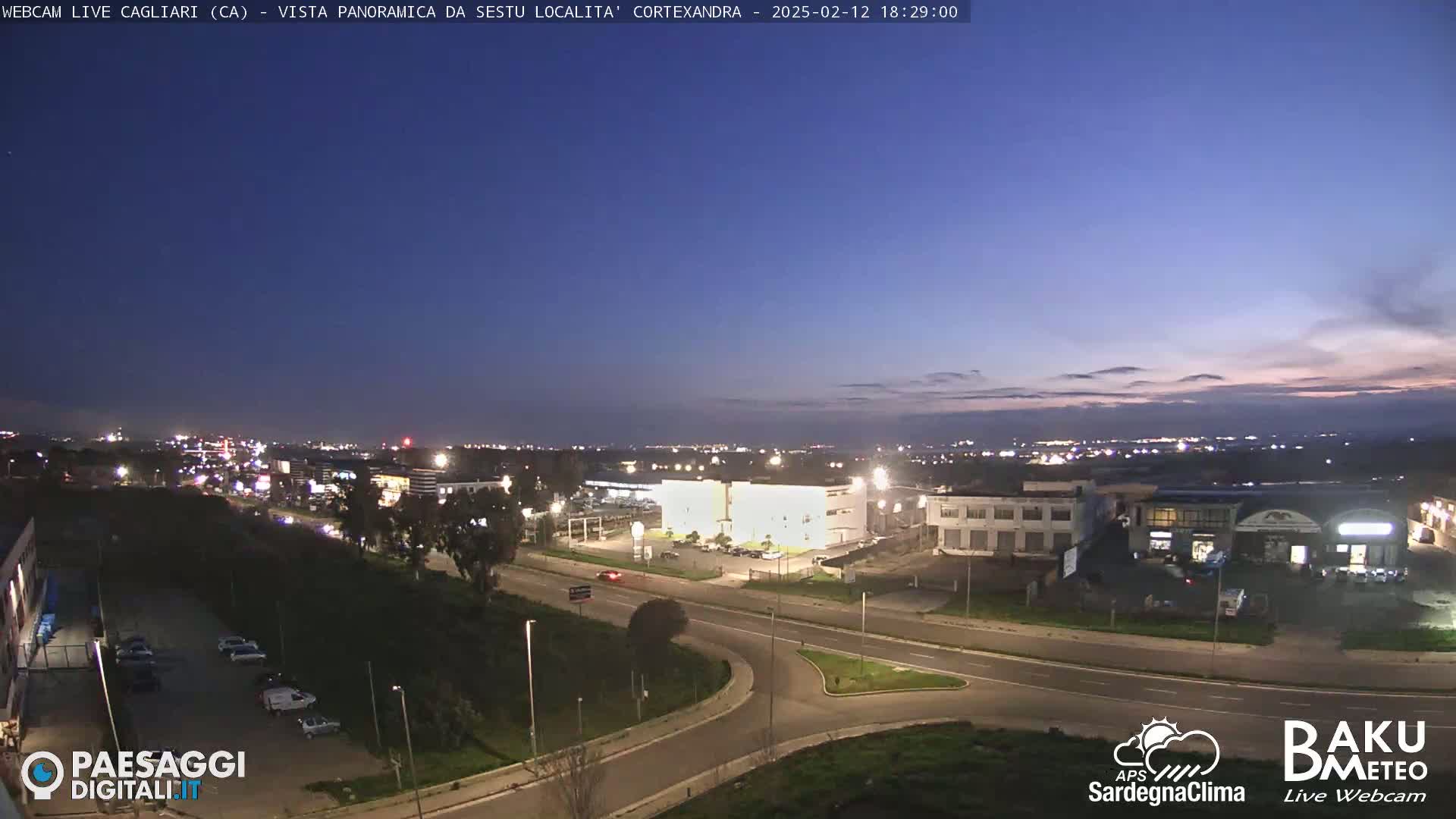 A twilight view of a city's outskirts, showing roads, buildings, and parked cars under a mostly clear, dark blue sky.
