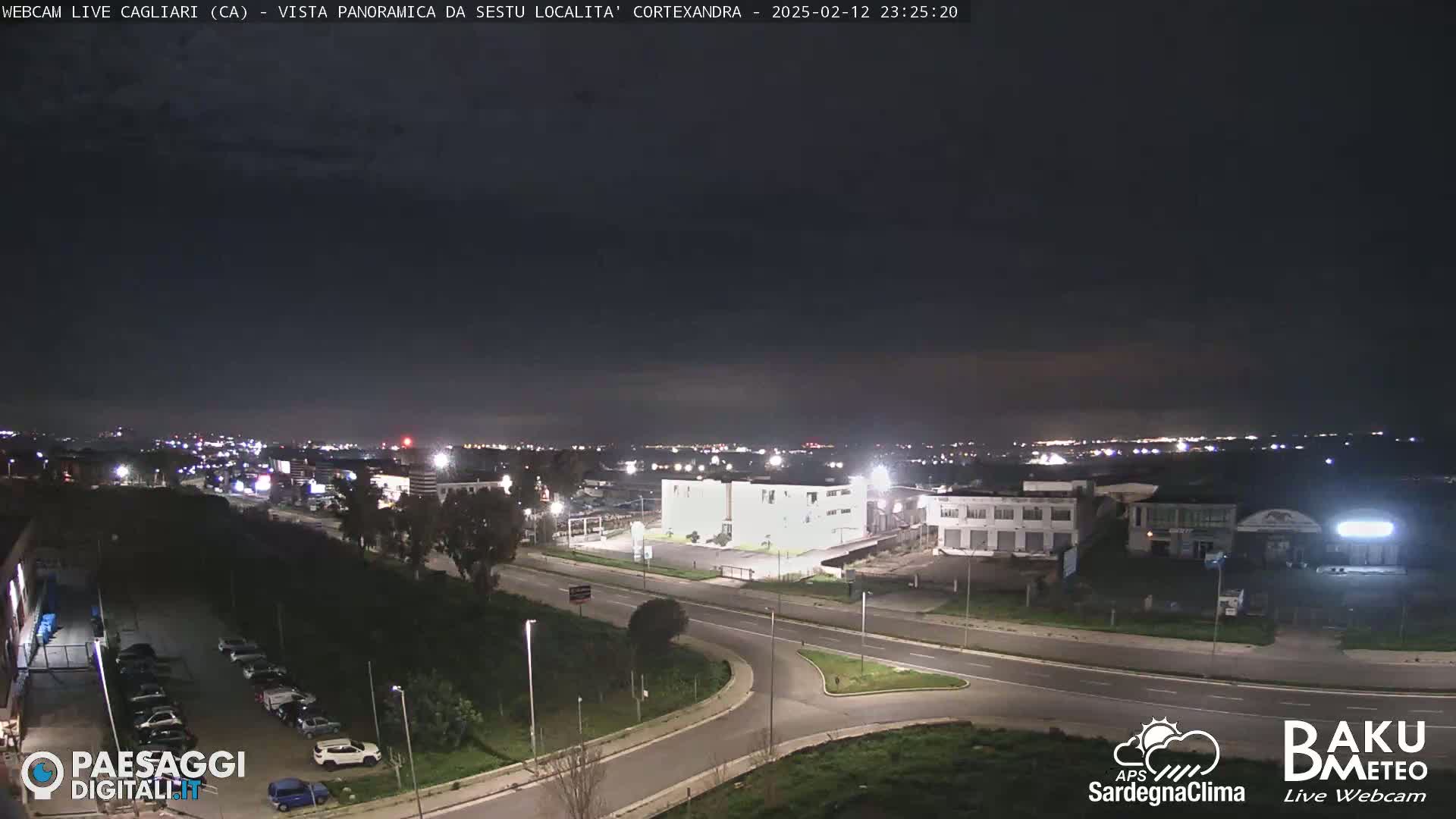 A nighttime, long shot of a road intersection and industrial area, with city lights visible in the distance under a clear, dark sky.
