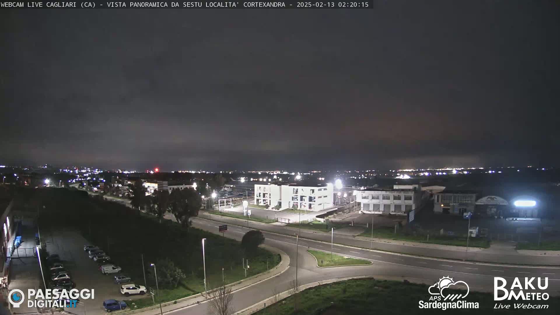 A nighttime, panoramic view of a city showing roads, buildings, and a few parked cars under a dark, clear sky.
