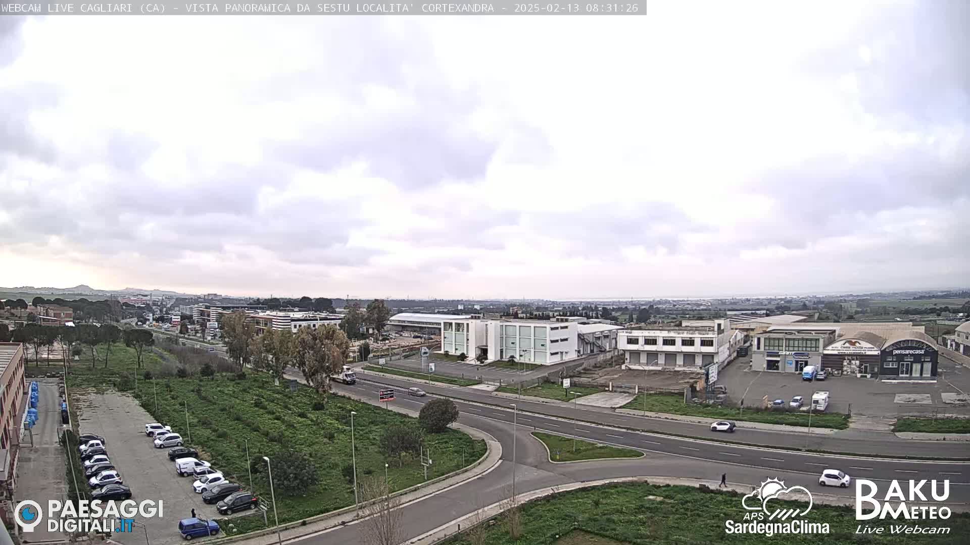 A panoramic view of a city with overcast skies shows a road intersection surrounded by buildings, parking lots, and green areas.