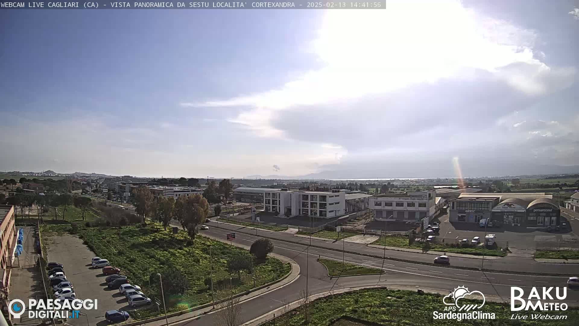 A mostly sunny day reveals a panoramic view of a city with industrial buildings, roads, and parked cars, under a partly cloudy sky.