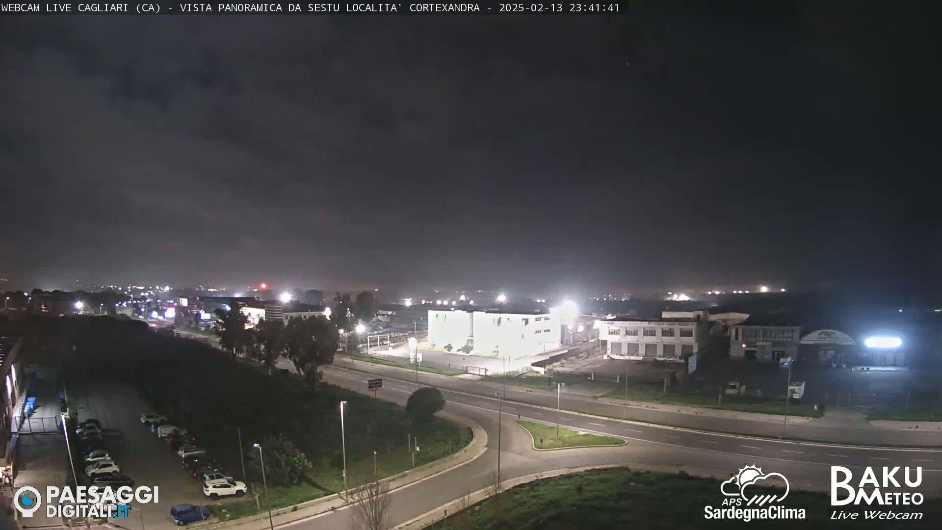 A nighttime, overcast view of a road intersection and industrial area with cars parked in lots and some lights illuminating buildings.