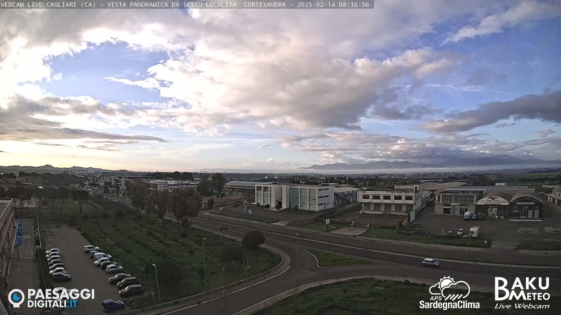 A partly cloudy day reveals a panoramic view of a city with industrial buildings and a road system, mountains visible in the distance.