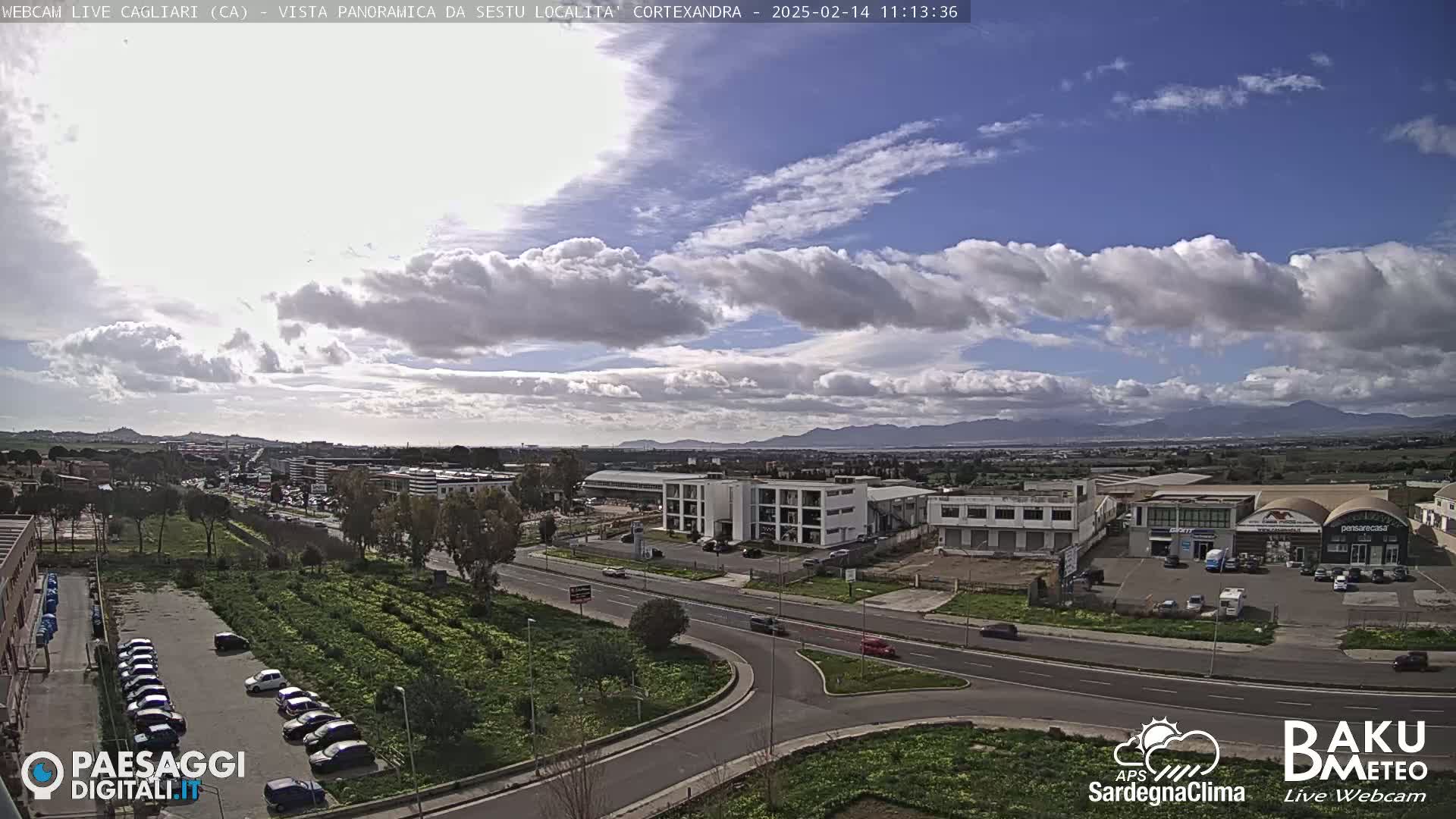 A partly cloudy day reveals a view of a city with industrial buildings, roads, and parking lots, and distant mountains under a mostly blue sky.