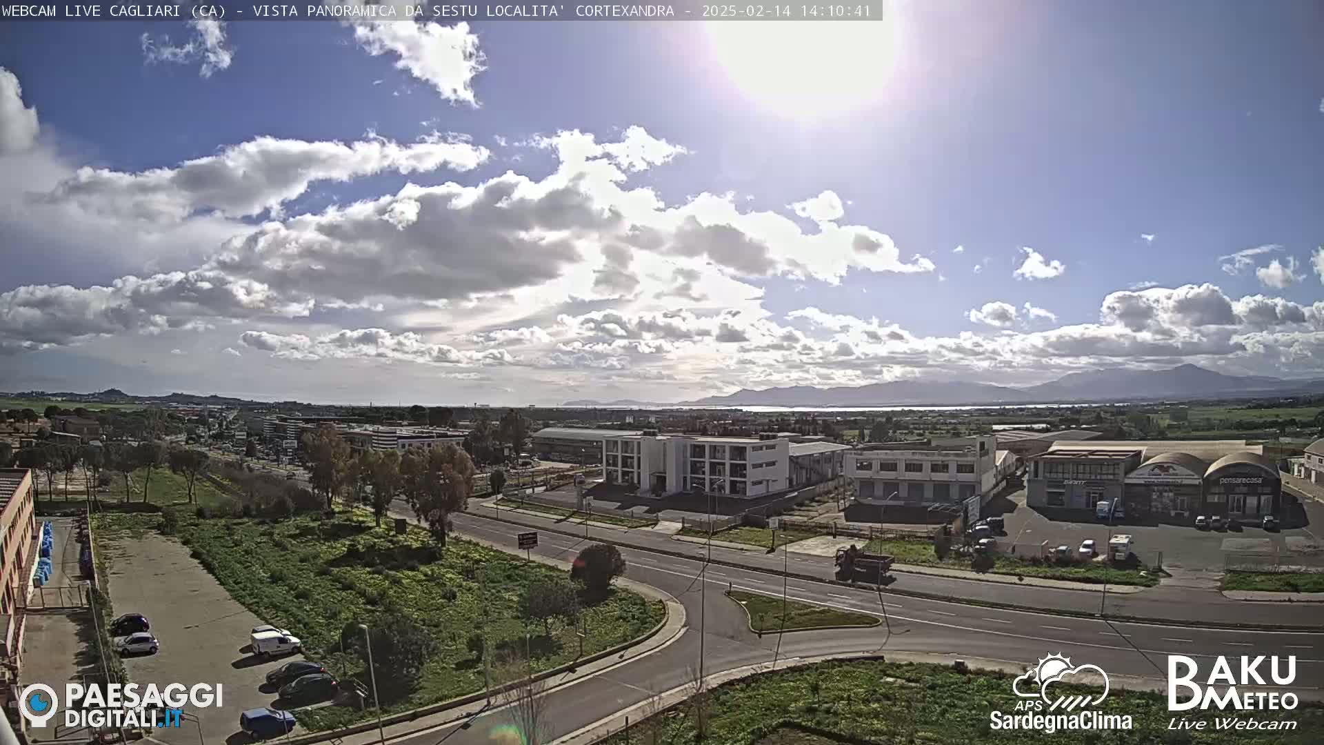 A partly sunny day reveals a panoramic view of a city with industrial buildings and roads, mountains and a body of water visible in the distance.