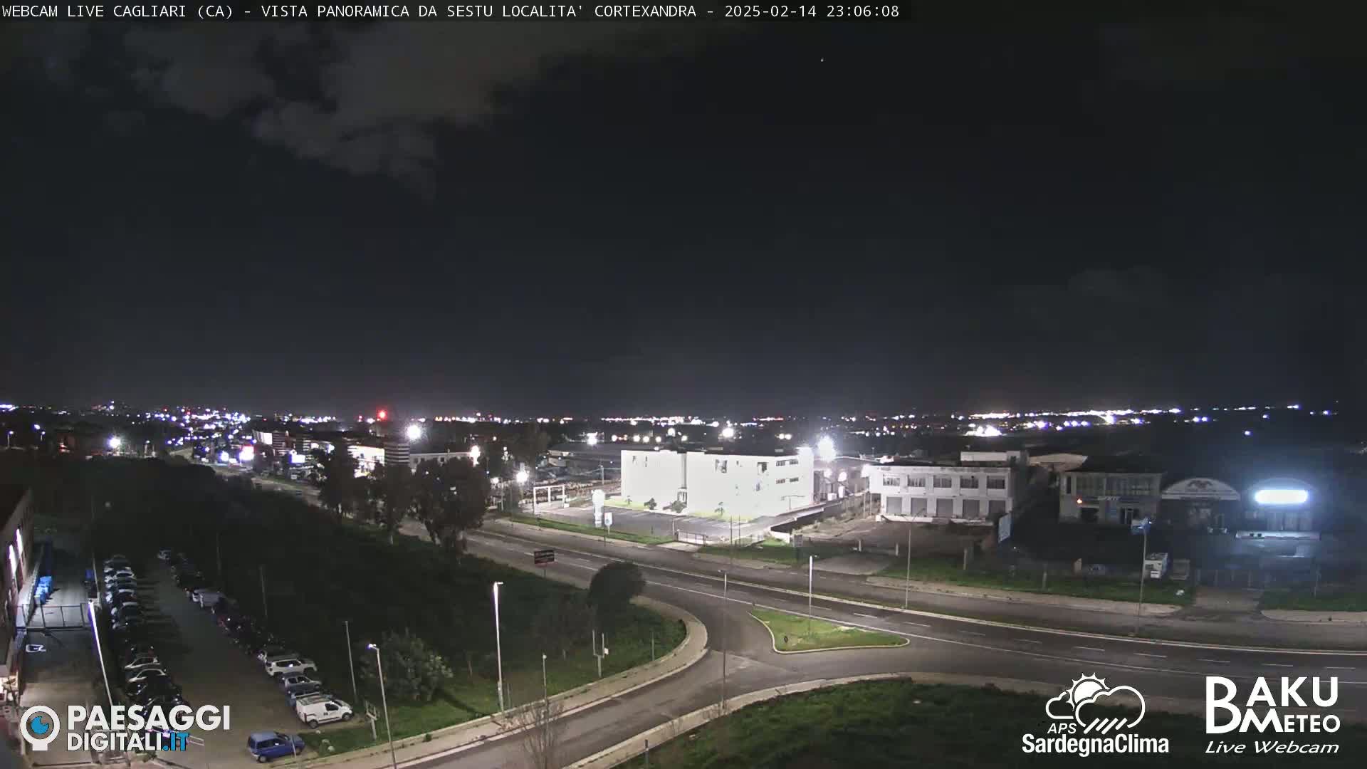 A nighttime, partly cloudy view of a city road intersection and surrounding buildings, with distant city lights visible in the background.