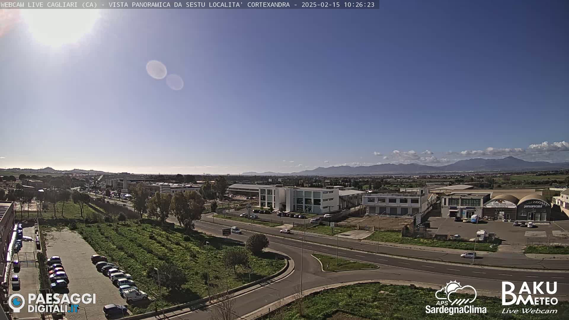 A sunny day reveals a panoramic view of a city with industrial buildings, roads, and parked cars, mountains visible in the distance.