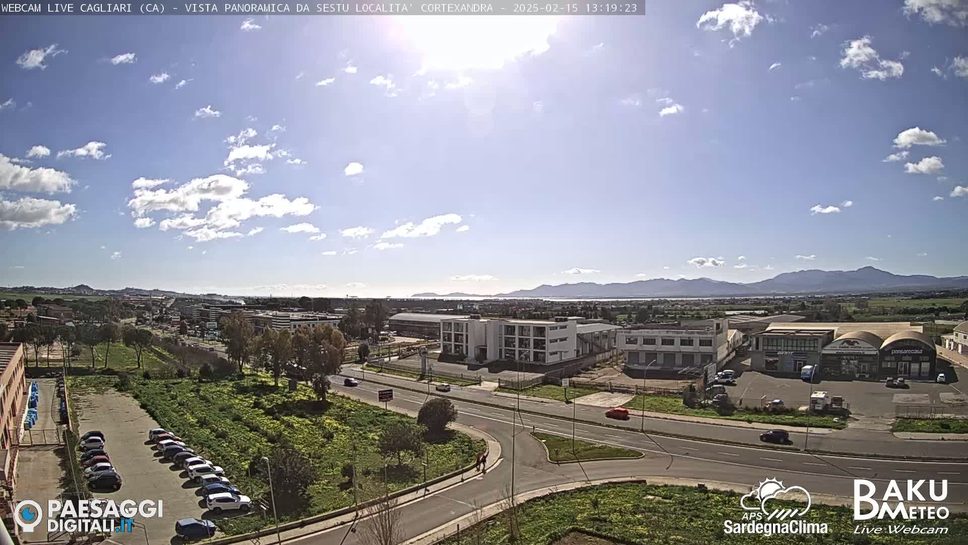 A sunny day with scattered clouds reveals a panoramic view of a city with industrial buildings, roads, and parked cars, extending to distant mountains.