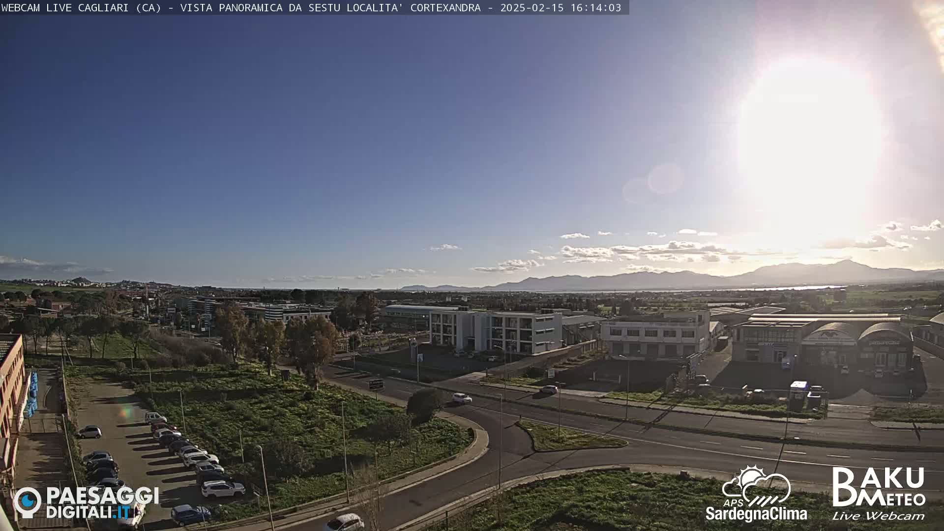 A sunny day reveals a panoramic view of a city with roads, buildings, and a parking lot, all situated in front of distant mountains.