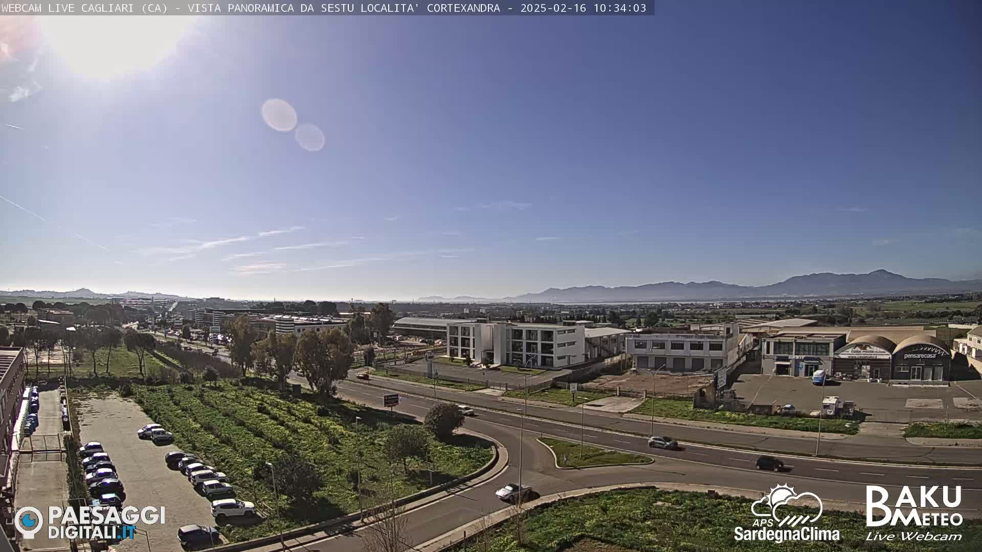A sunny, clear day overlooks a highway intersection, industrial buildings, and parking lots, with distant mountains visible on the horizon.