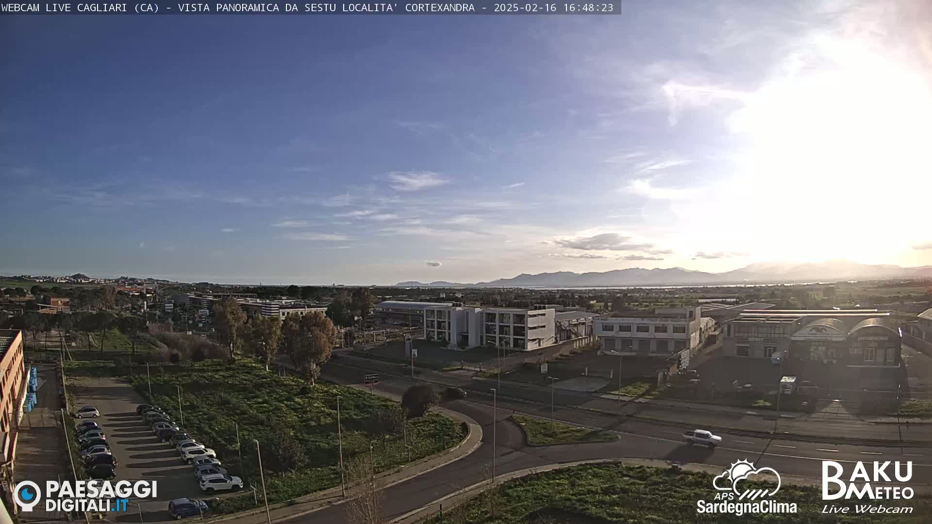 A mostly sunny day reveals a panoramic view of a city with industrial buildings and a parking lot, extending to distant mountains under a partly cloudy sky.