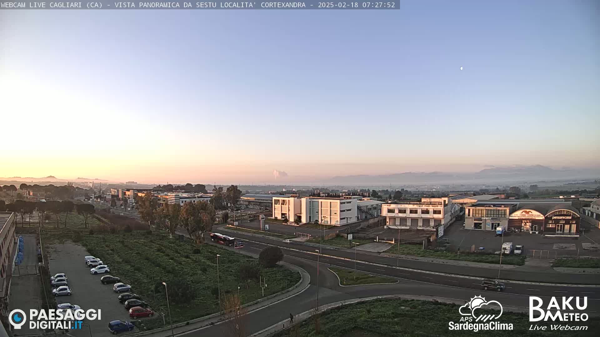 A hazy sunrise illuminates a flat landscape of industrial buildings, roads, and parked cars, with distant mountains barely visible in the background.