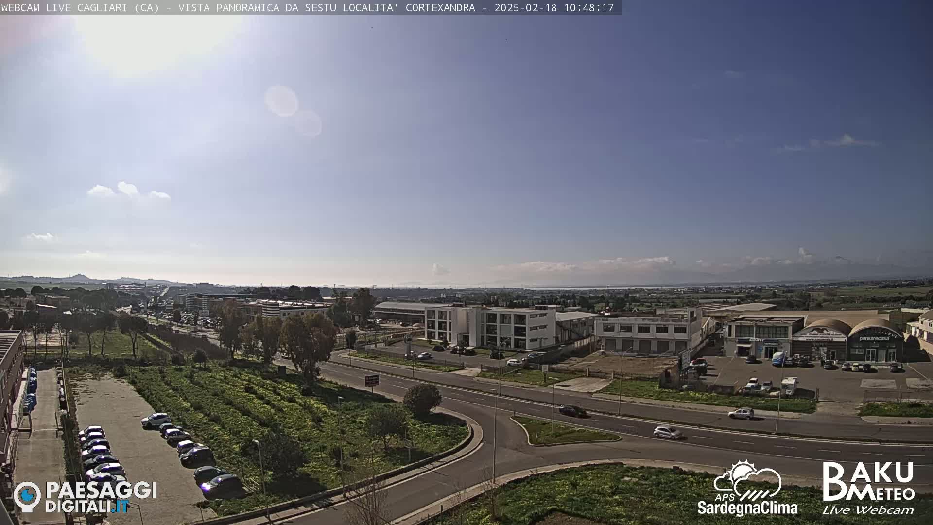 A sunny, mostly clear day reveals a panoramic view of a city's outskirts, featuring industrial buildings, roadways, and a parking lot filled with cars, with distant mountains visible on the horizon.