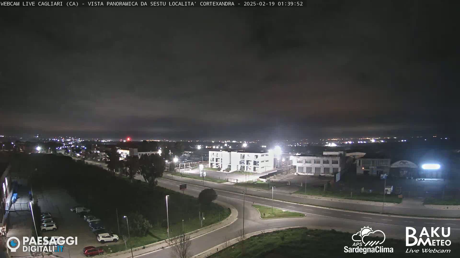 A nighttime, slightly overcast view of a road intersection and industrial area, with city lights visible in the distance.
