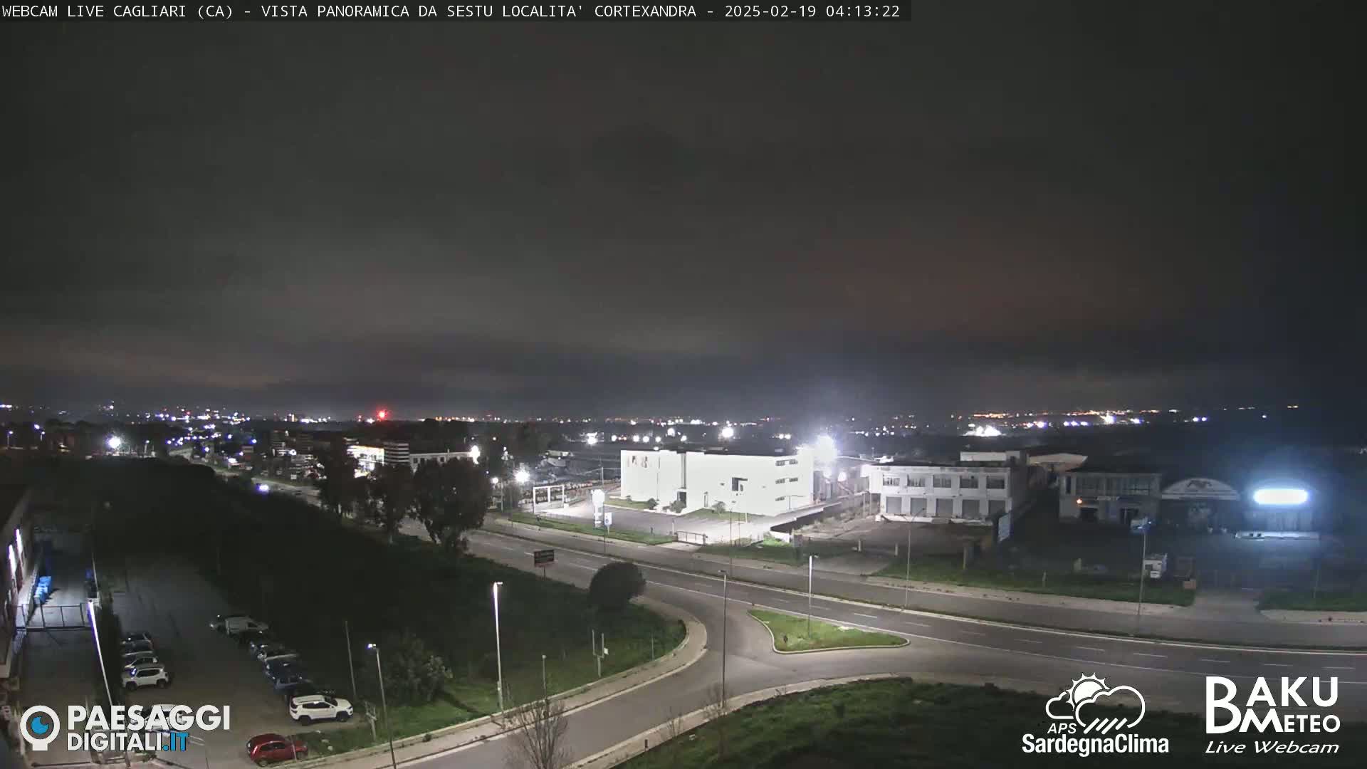 A nighttime, overcast view of a road intersection and industrial area, with distant city lights visible on the horizon.
