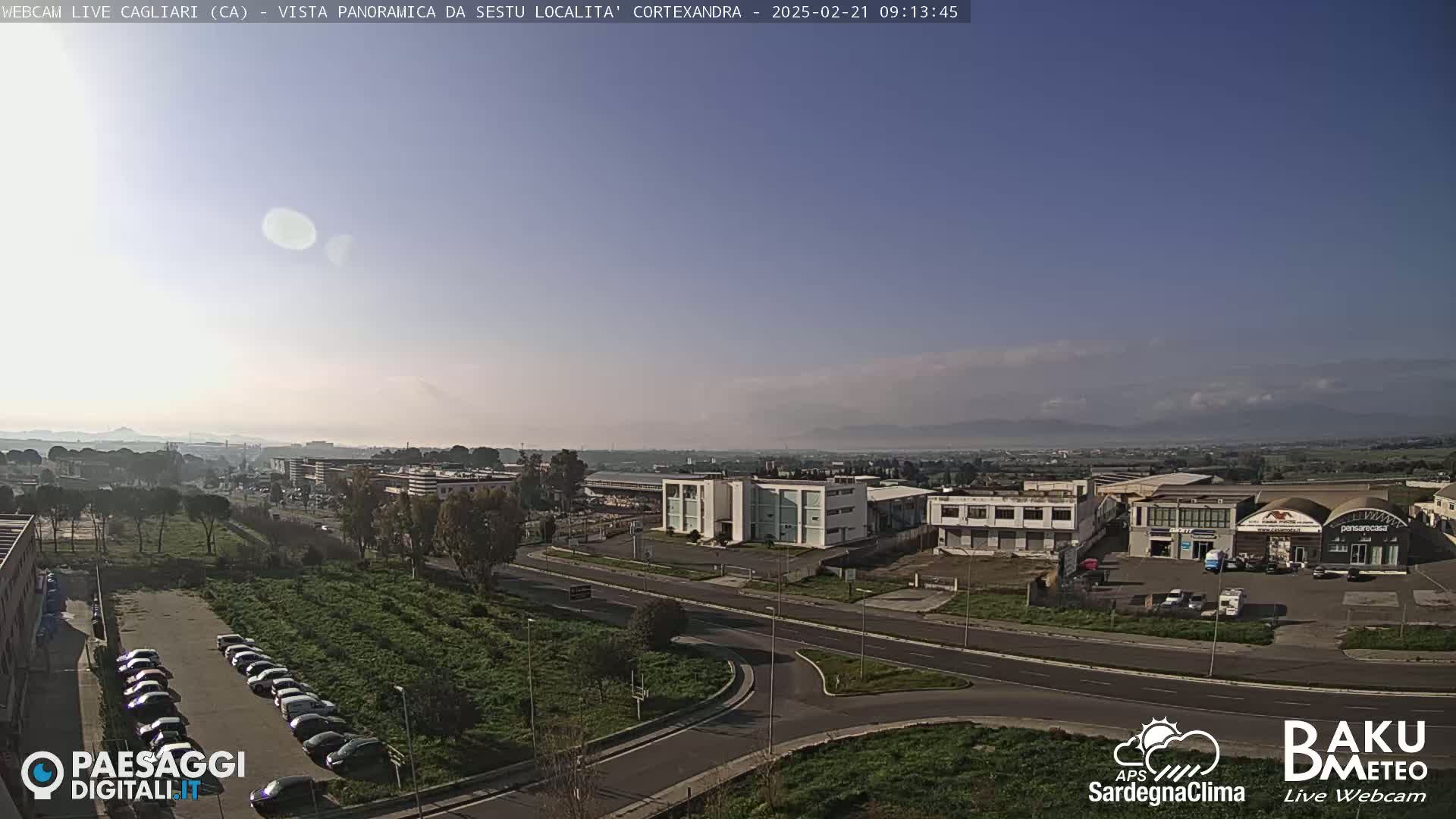 A mostly clear, bright day reveals a panoramic view of a city's outskirts, featuring industrial buildings, roads, parked cars, and distant mountains under a pale blue sky.