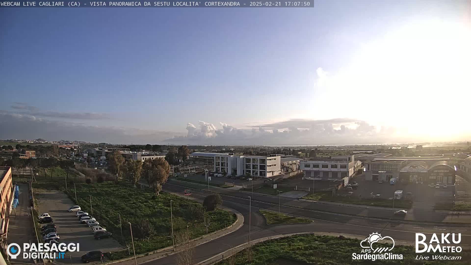 A mostly clear, sunny day reveals a panoramic view of a city, featuring industrial buildings, roads, and parked cars, with a band of clouds on the horizon.
