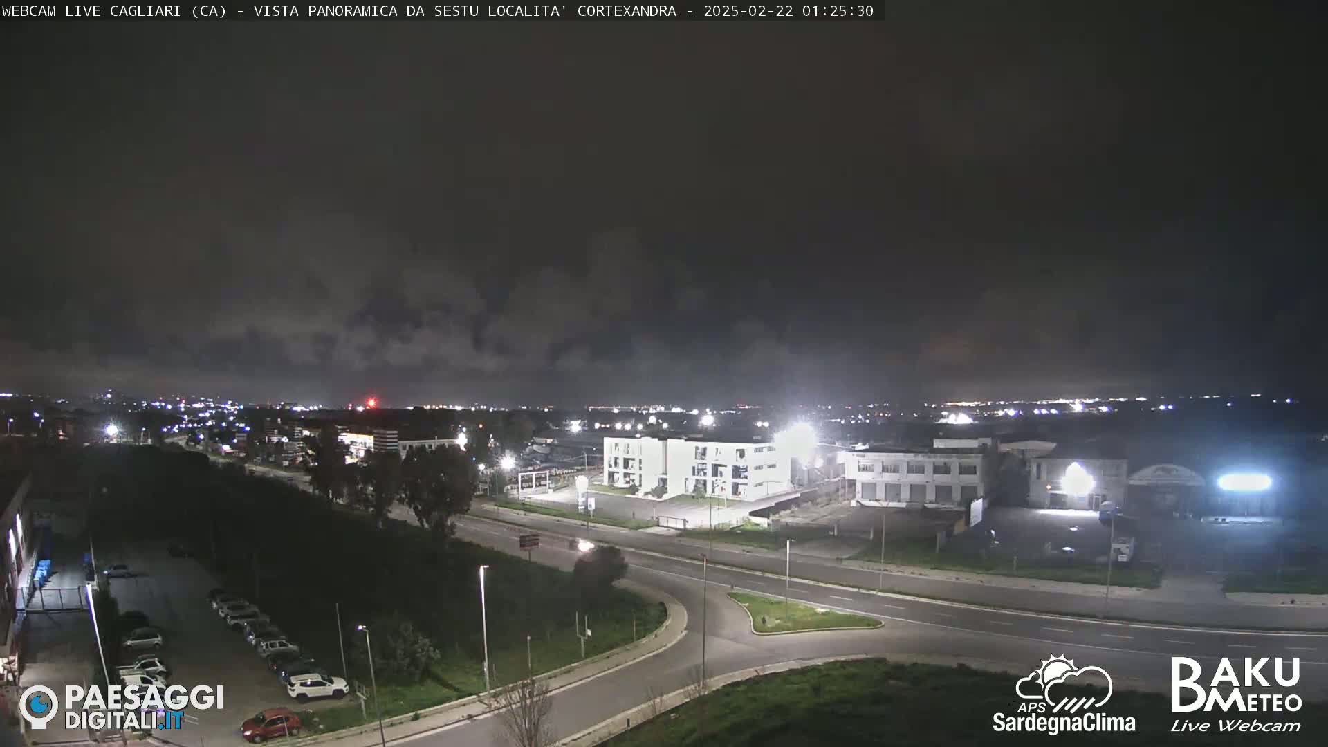 A nighttime view of a city with overcast skies, showing roads, buildings, and parked cars.