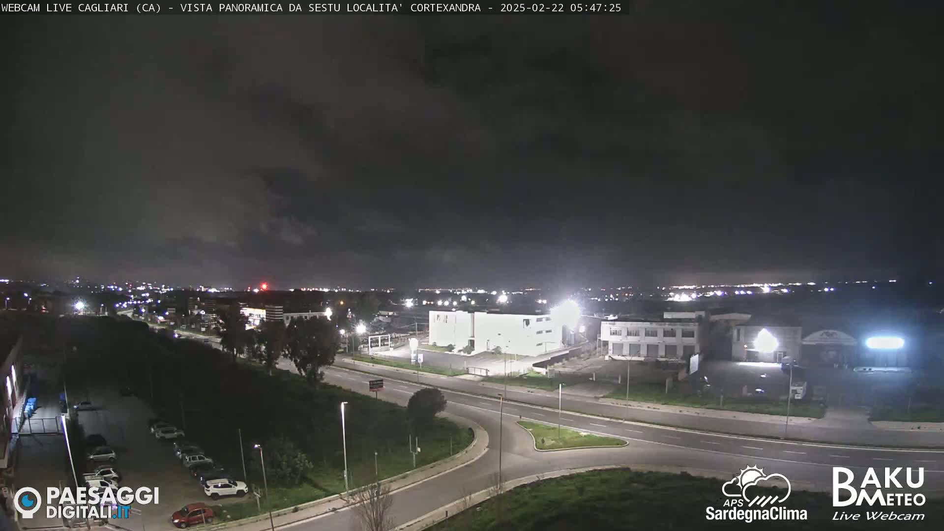 A nighttime, mostly clear view of a city's outskirts, featuring roads, buildings, and parked cars.