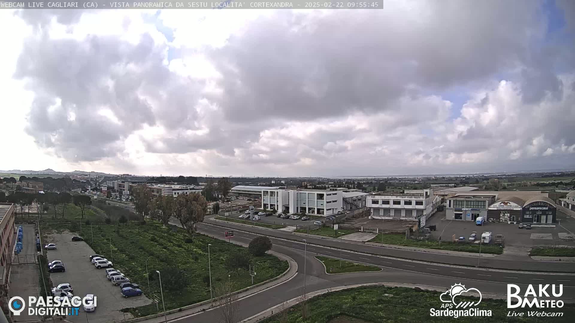 A panoramic view of a city under a mostly cloudy sky shows roads, buildings, and parked cars.