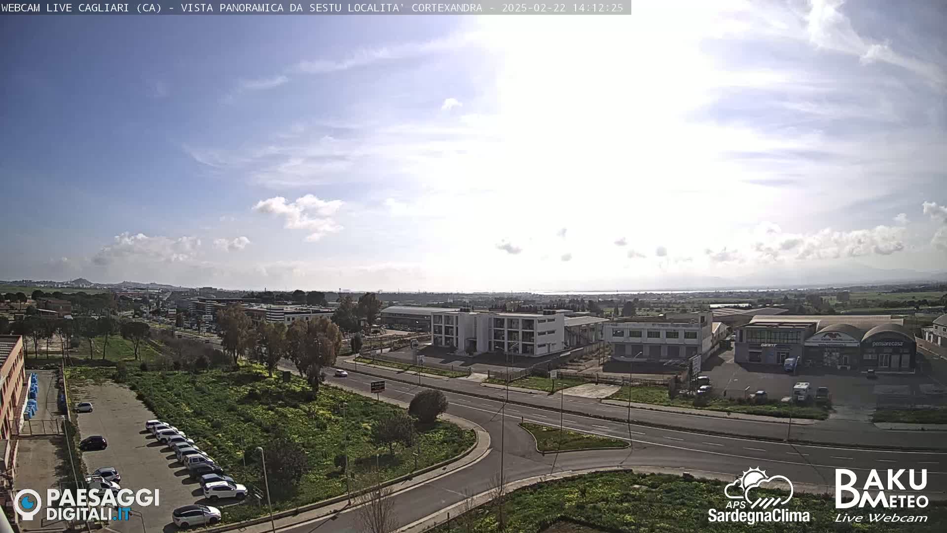 A partly sunny day reveals a panoramic view of a city with industrial buildings and roadways, featuring a parking lot filled with cars and light green vegetation.