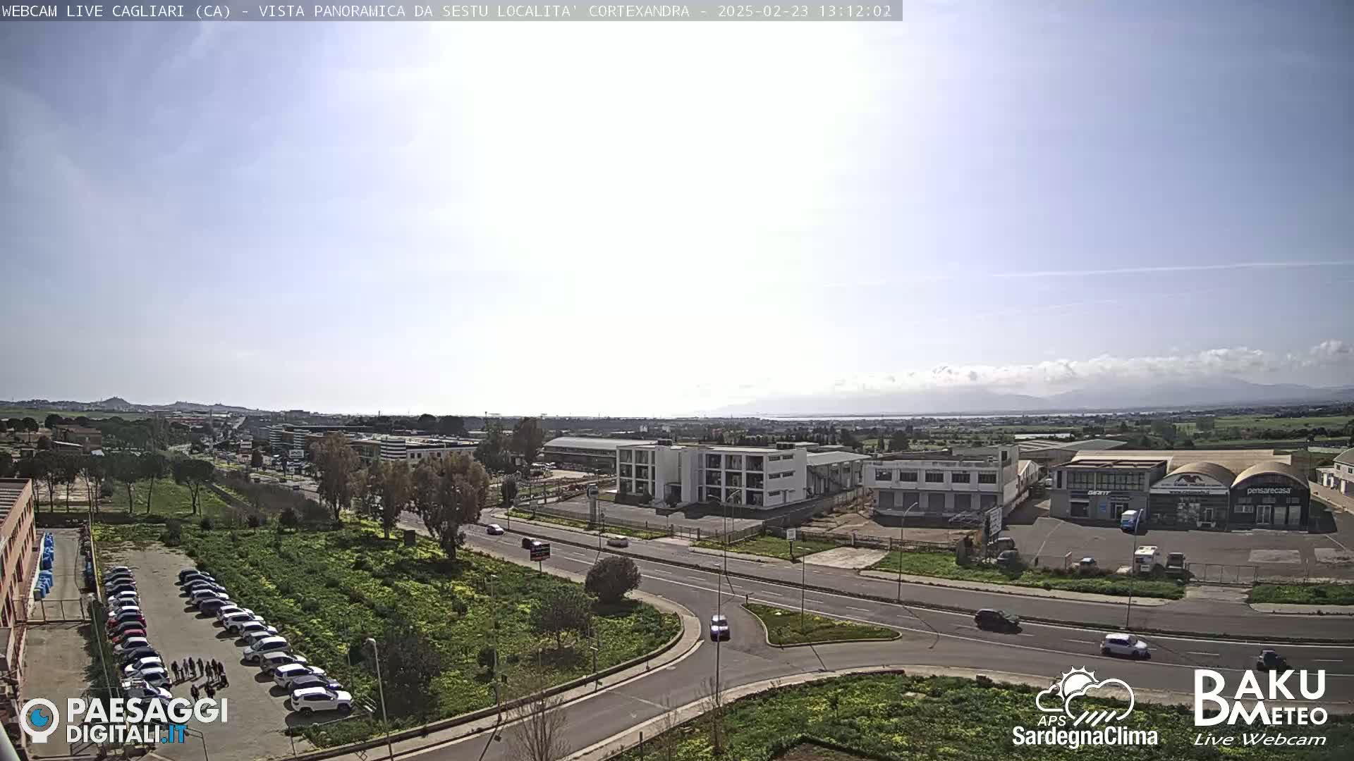 A sunny day reveals a panoramic view of a city with roads, buildings, and parked cars, extending to a distant mountain range and body of water under a mostly clear sky with some scattered clouds.